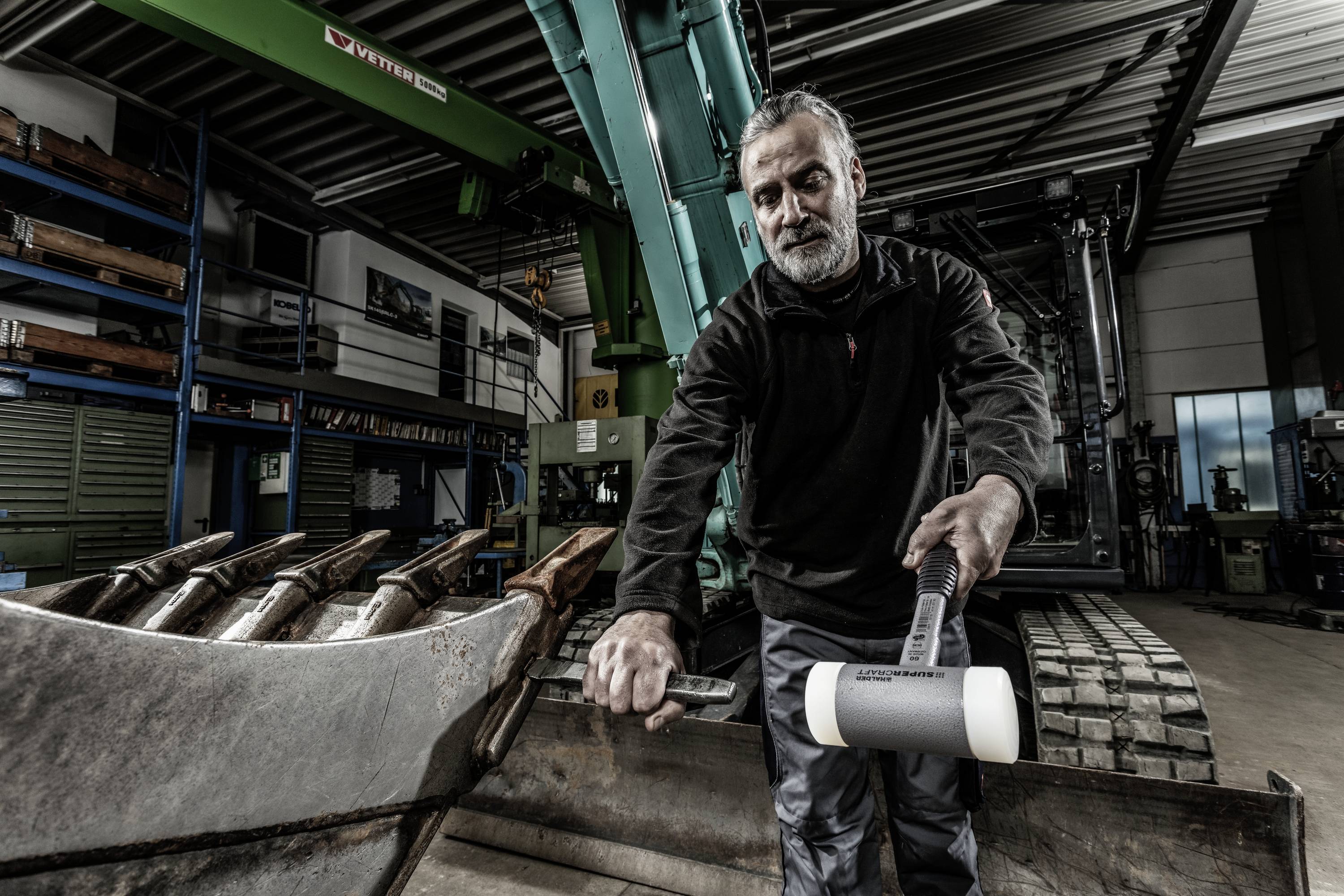 Mężczyzna w warsztacie pracuje przy dużej łyżce koparki, trzymając w ręku młotek. W tle widoczne są regały i maszyny.