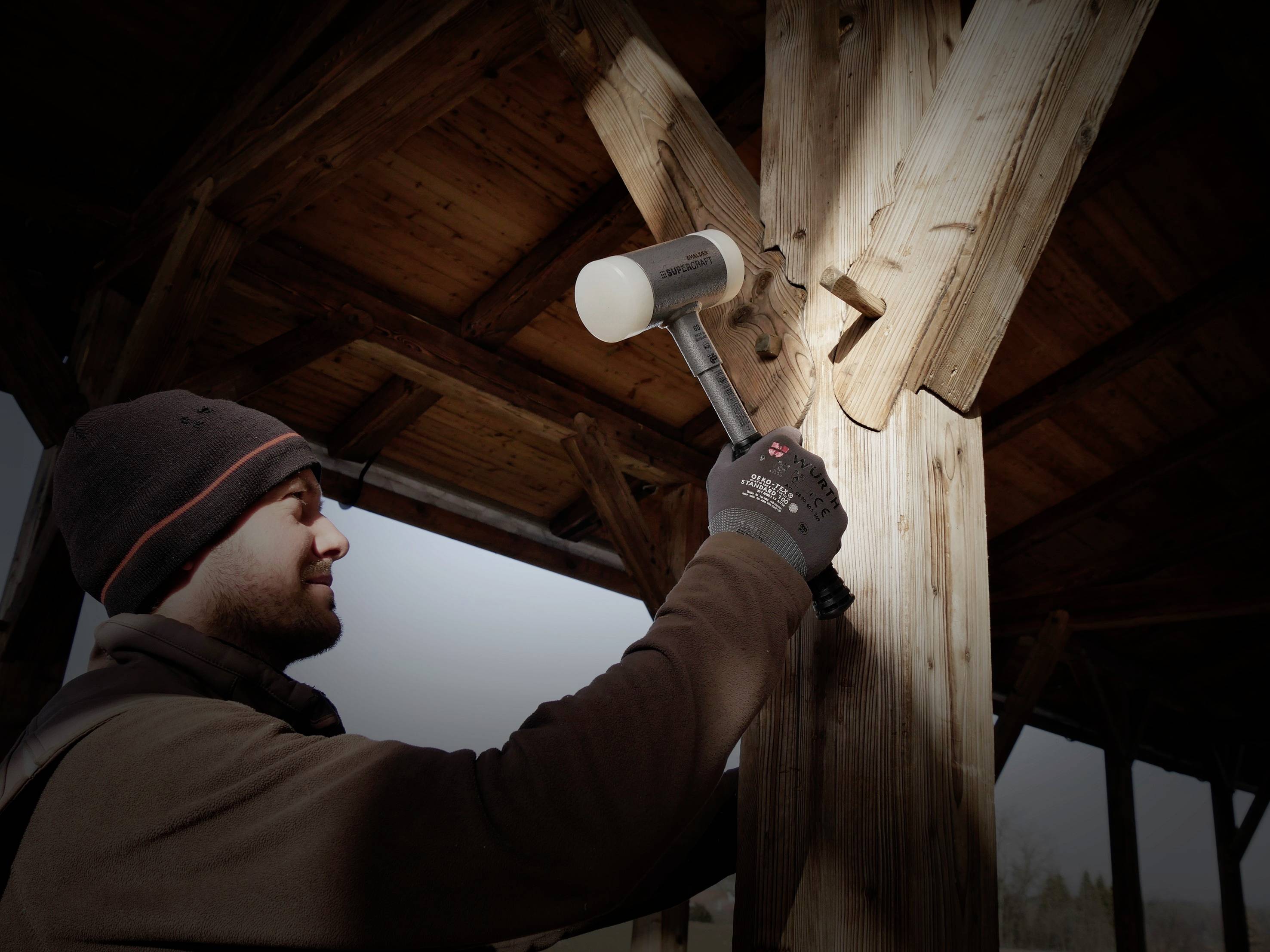 Rzemieślnik uderza gumowym młotkiem w drewniany belk w zadaszonym pomieszczeniu. Mężczyzna ma na sobie rękawice i czapkę.
