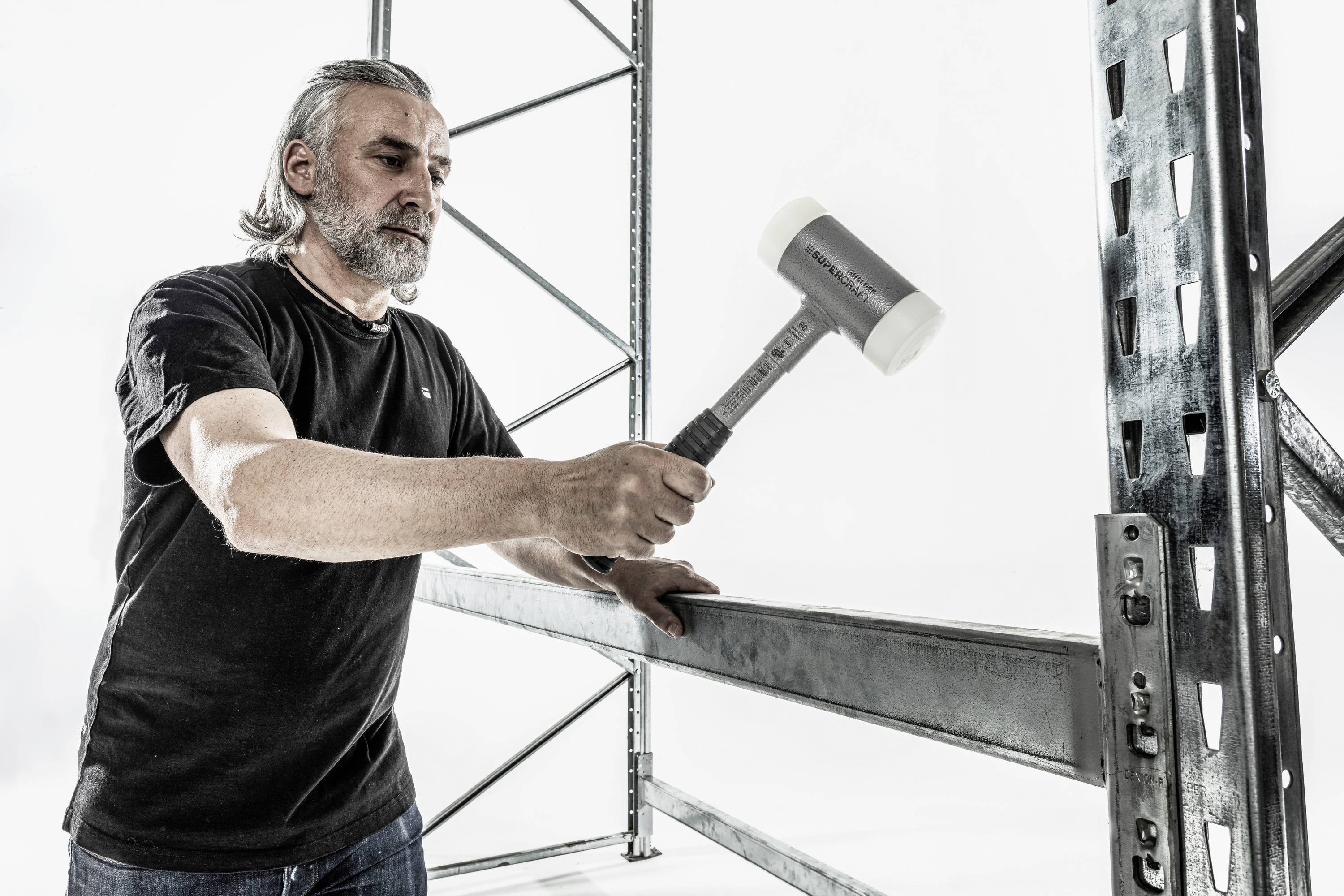 Mężczyzna używa młotka gumowego do montażu regału metalowego. Ma na sobie czarną koszulkę. W tle widoczna jest rama regału.