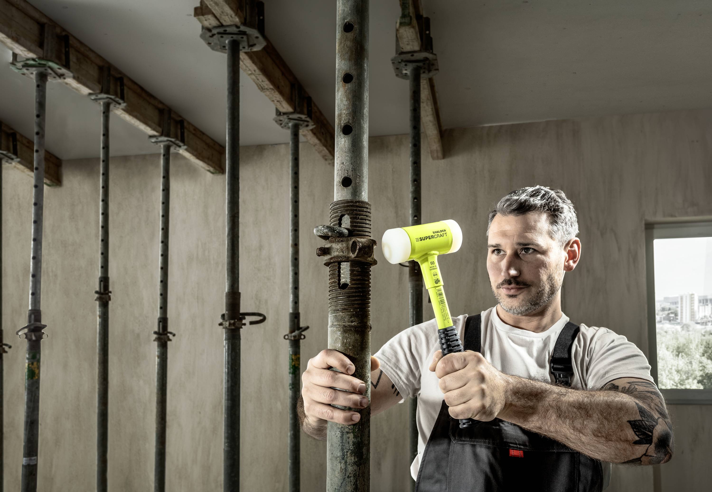 Robotnik budowlany uderza żółtym młotkiem w metalowy słup, aby go zabezpieczyć. W tle widoczne są liczne podpory.