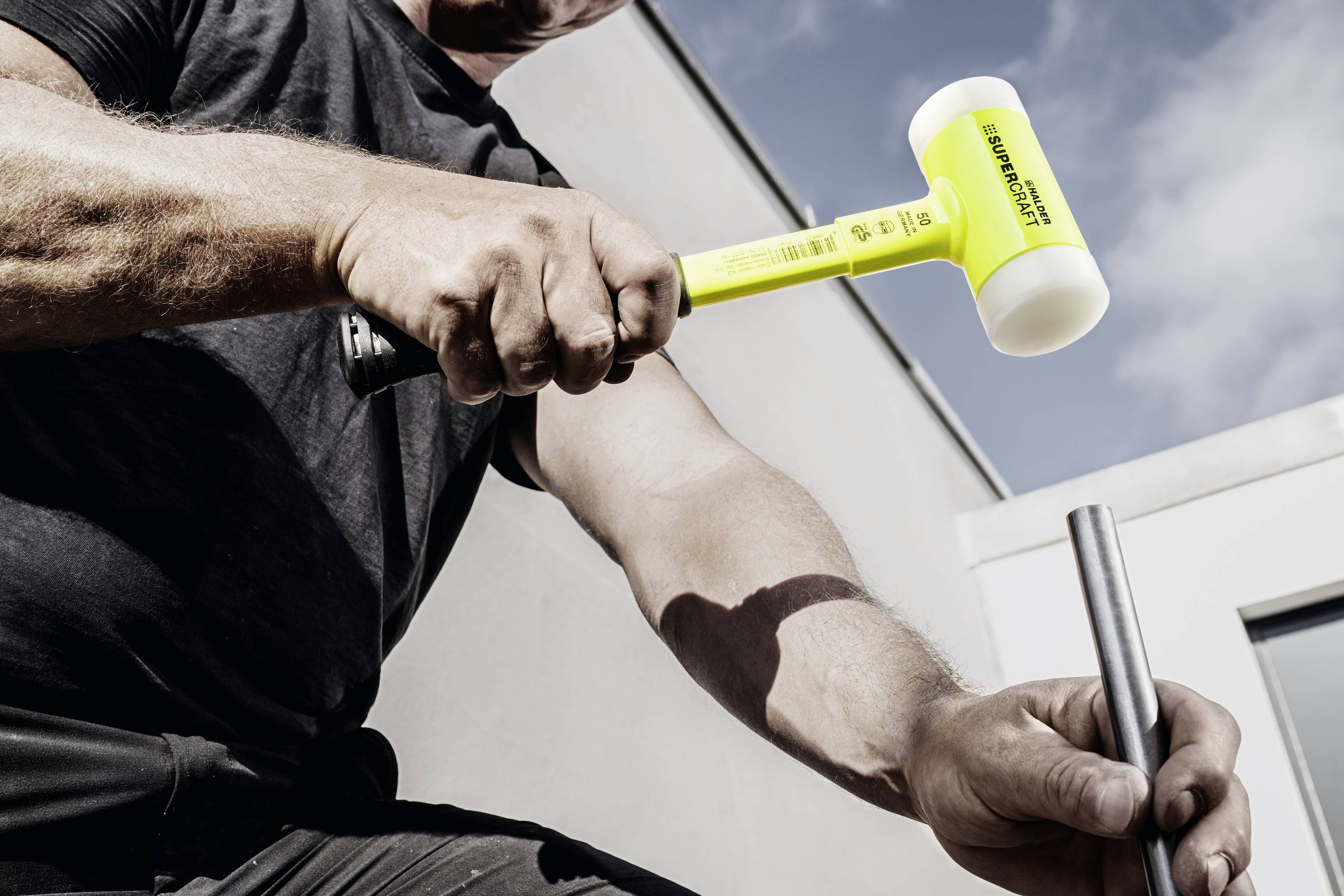 Mężczyzna wbija żółtym gumowym młotkiem w metalową rurę na wolnym powietrzu. Niebo jest częściowo zachmurzone.