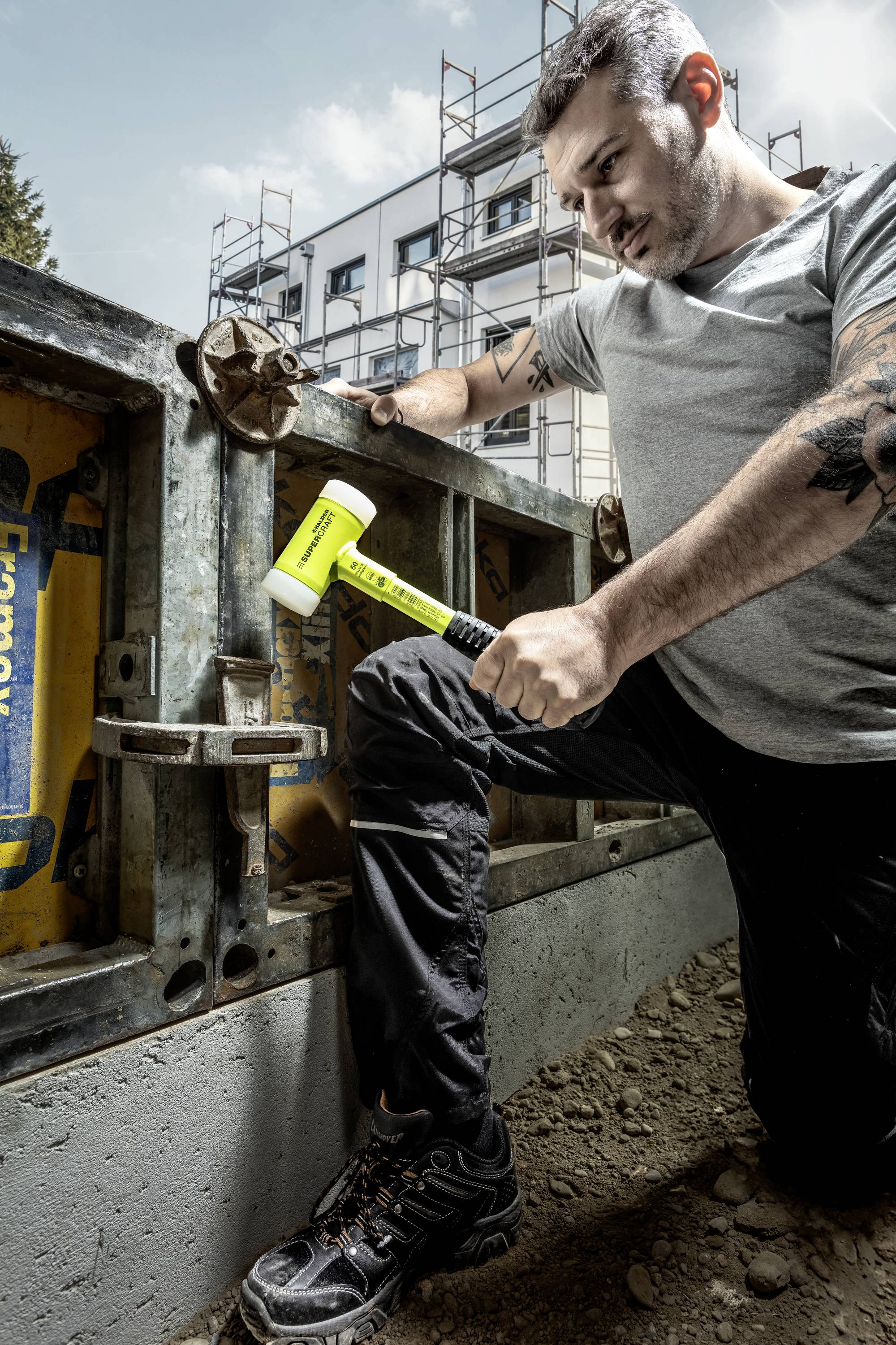 Mężczyzna w ubraniu roboczym klęczy na budowie i używa młotka do pracy przy szalunku. W tle widoczne są budynki i rusztowania.