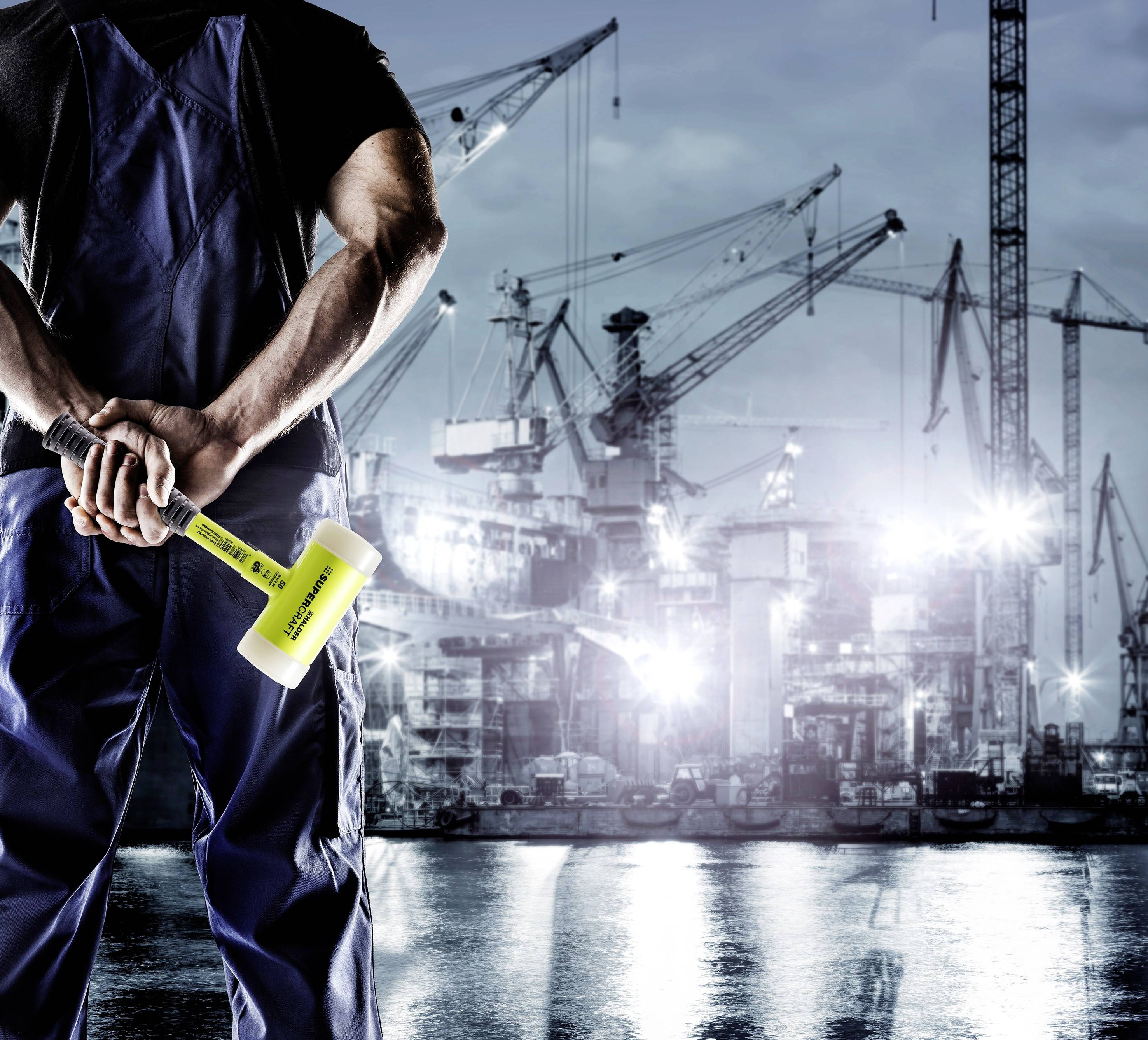 Robotnik w niebieskim kombinezonie roboczym trzyma duży młot przed stocznią z dźwigami w tle nocą.