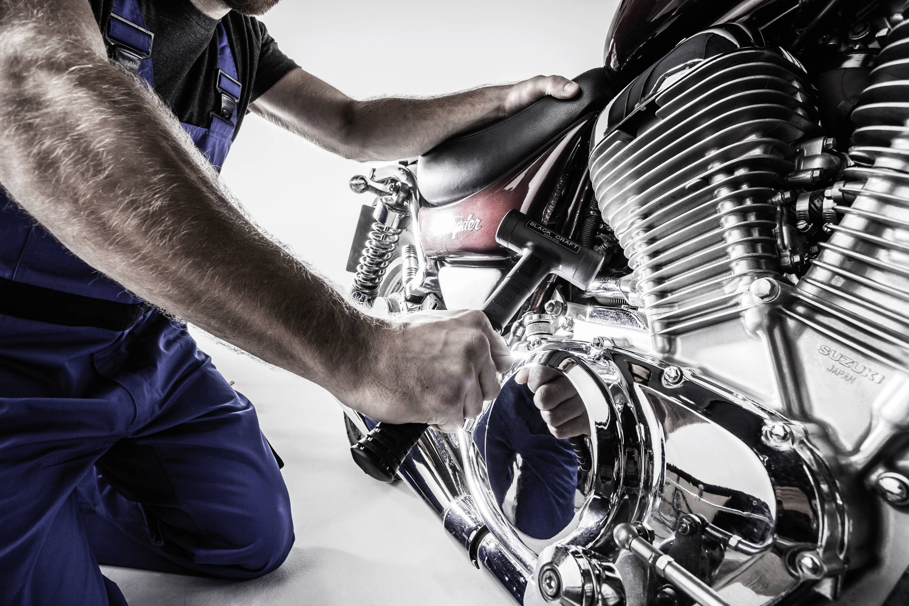 Osoba w odzieży roboczej naprawia motocykl, skupiając się na obszarze silnika.