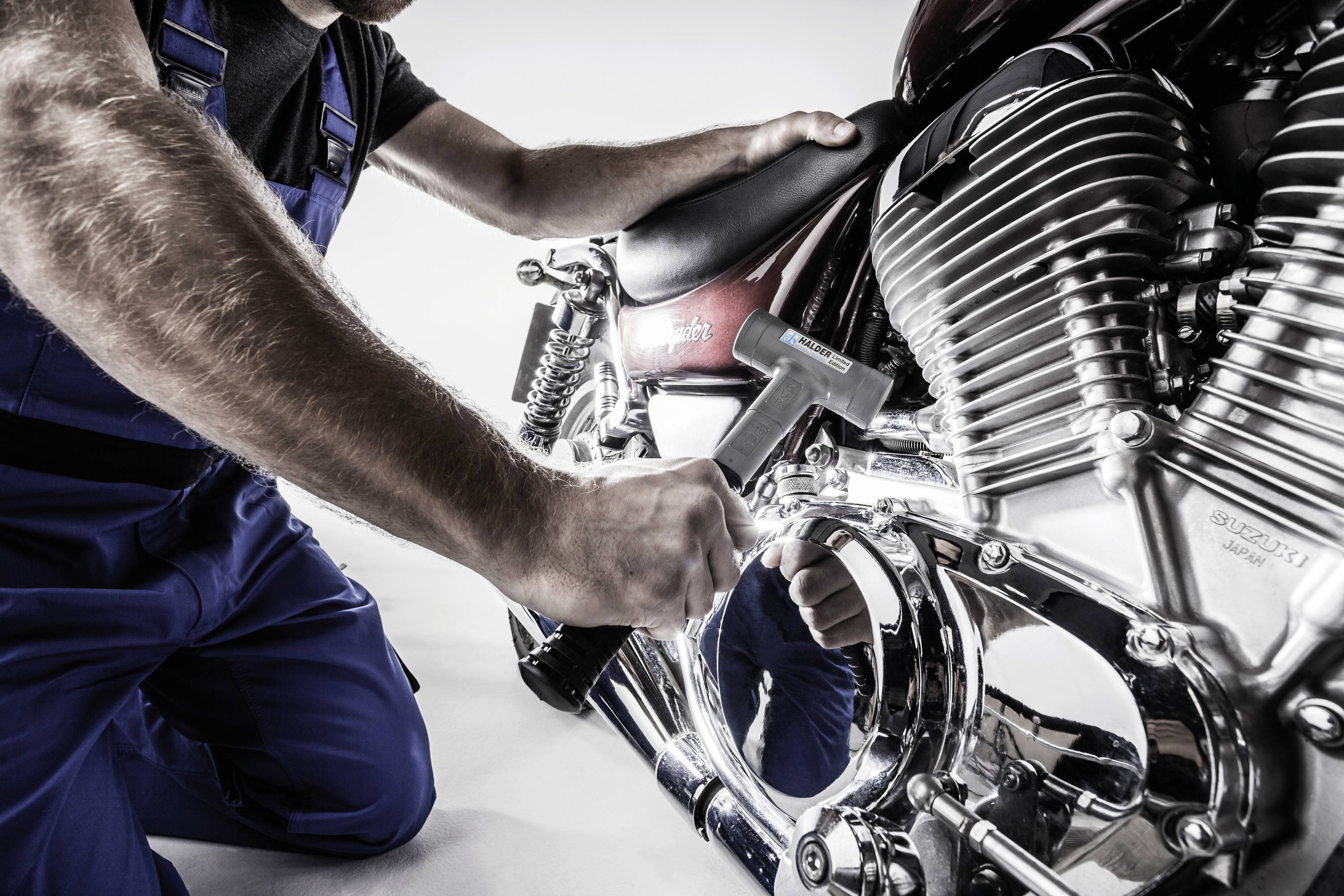 Osoba ubrana w niebieskie ogrodniczki pracuje z młotkiem przy motocyklu w jasnym pomieszczeniu.