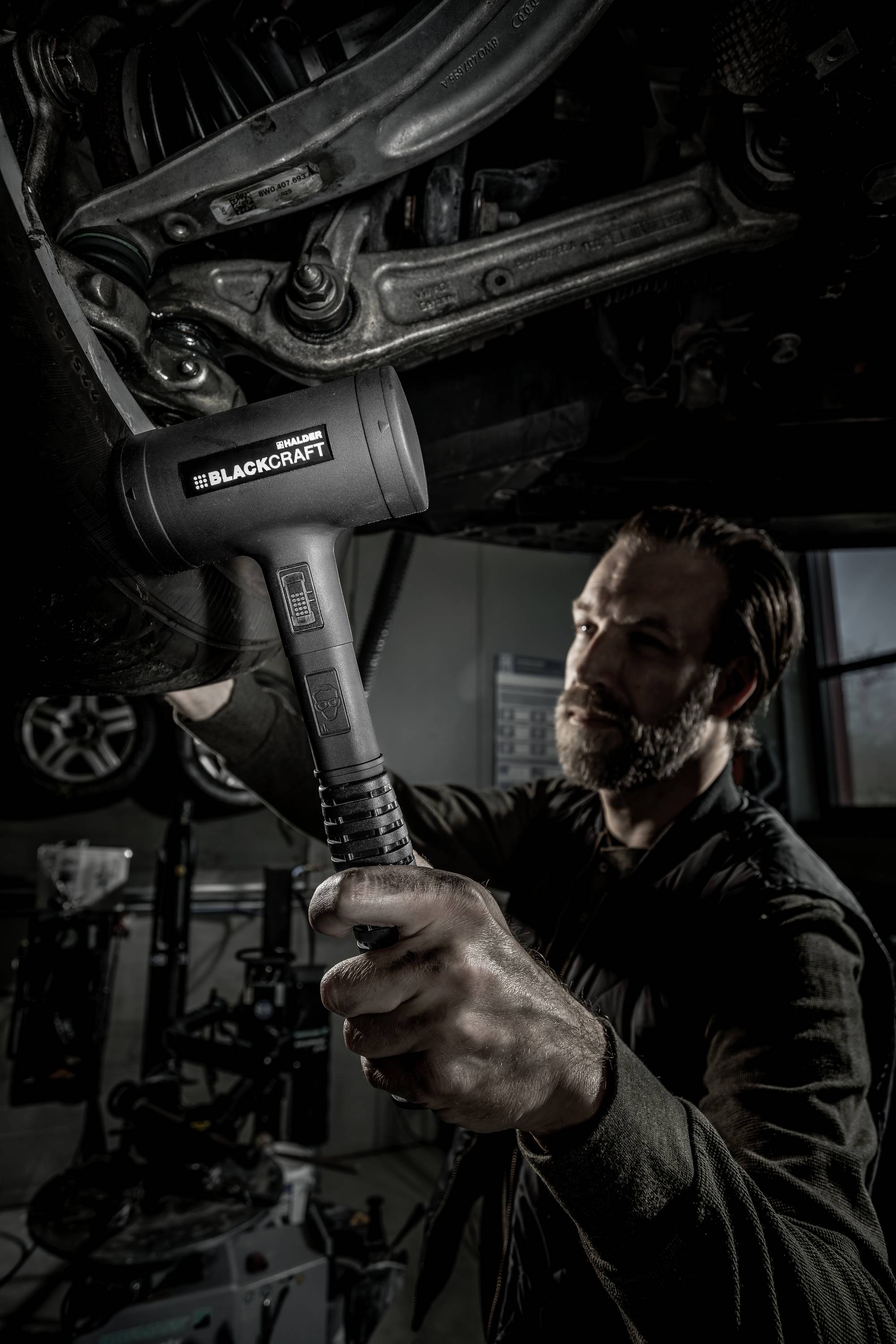 Mechanik pracuje w warsztacie pod samochodem i używa dużego gumowego młotka do pracy przy zawieszeniu.