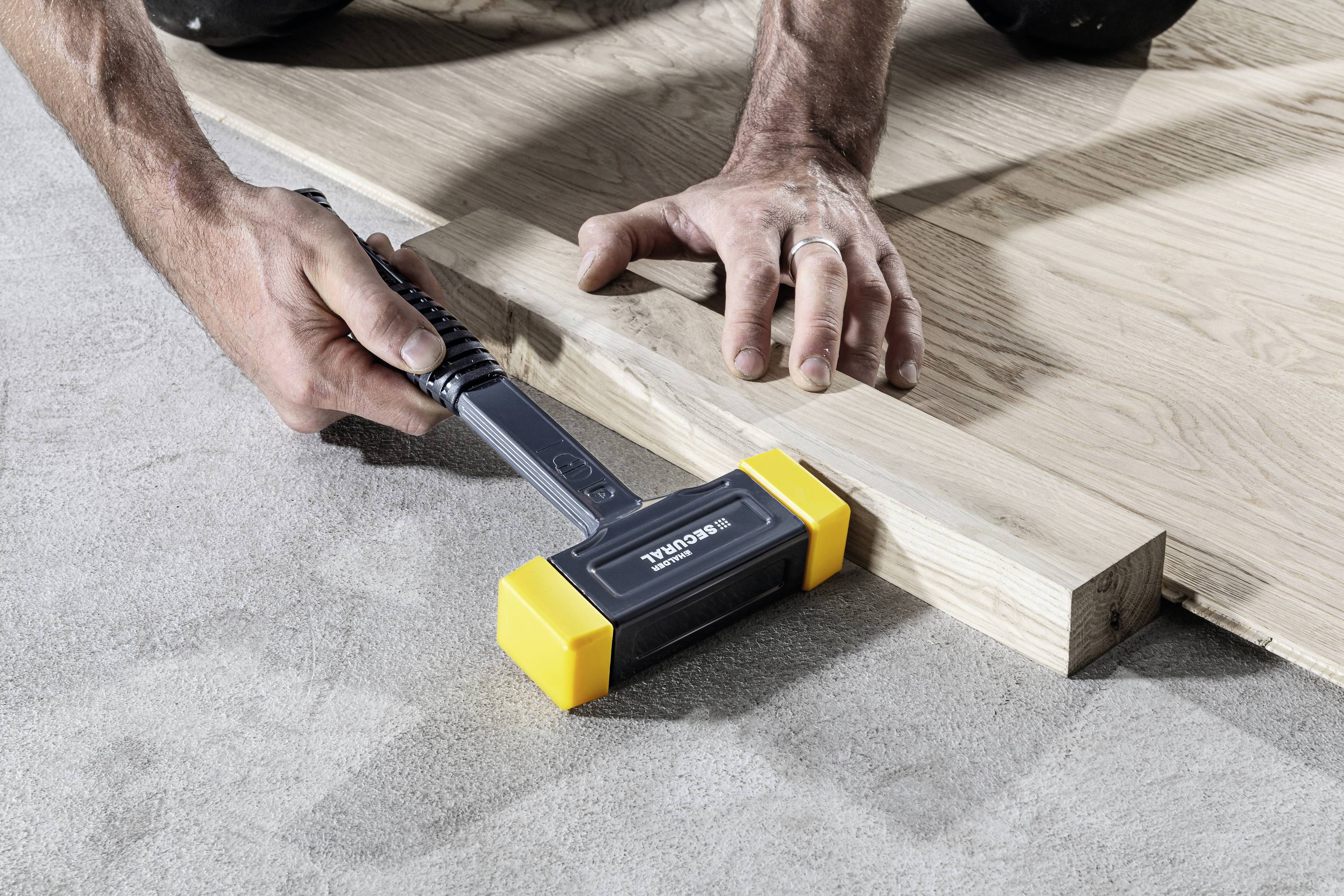 Osoba wbija czarnym narzędziem z żółtymi końcówkami drewnianą deskę w betonową podłogę. Ręce przytrzymują deskę.