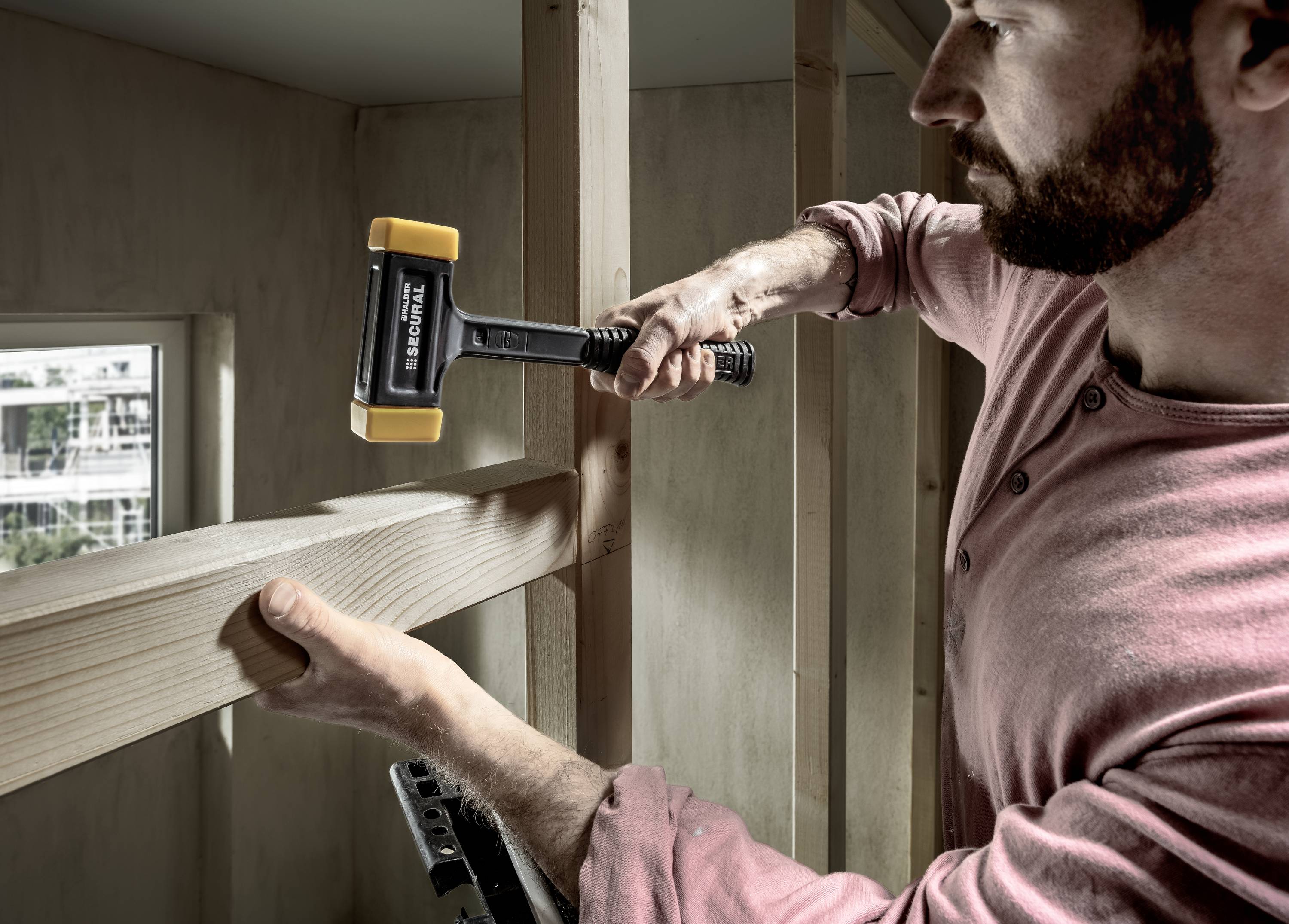 Mężczyzna wbija gwoździe w drewnianą deskę do konstrukcji ramowej. W tle widać okno z widokiem na budynek.