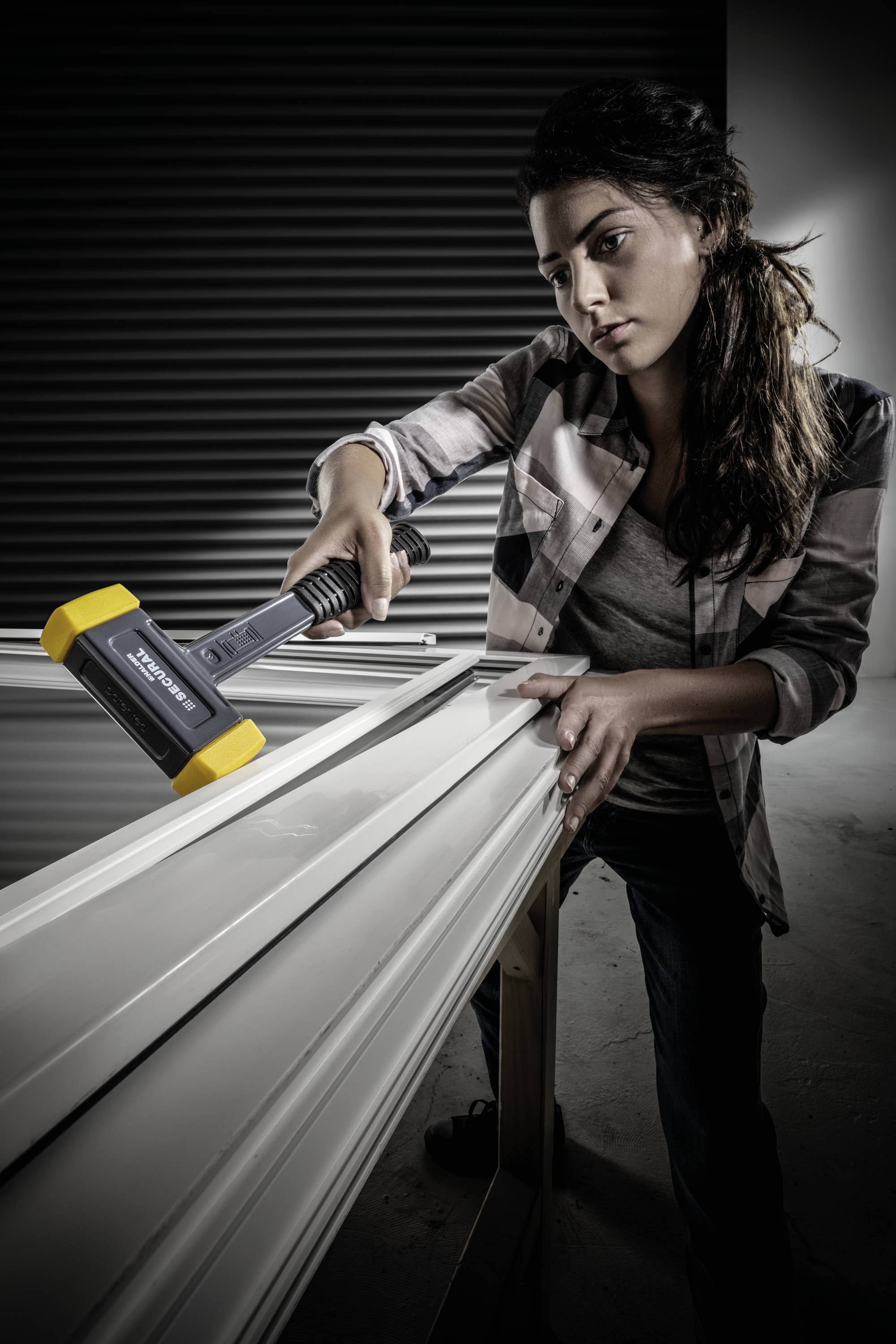 Osoba pracuje młotkiem przy framudze okiennej w warsztacie. Pomieszczenie jest słabo oświetlone.