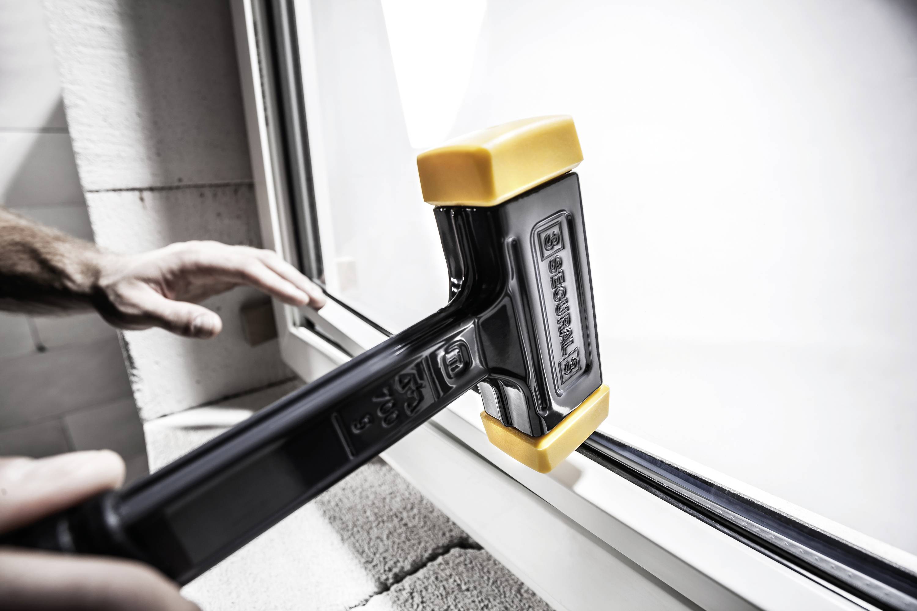 Osoba używa dużego młota z żółto-czarną głowicą do instalacji lub regulacji okna w budynku.