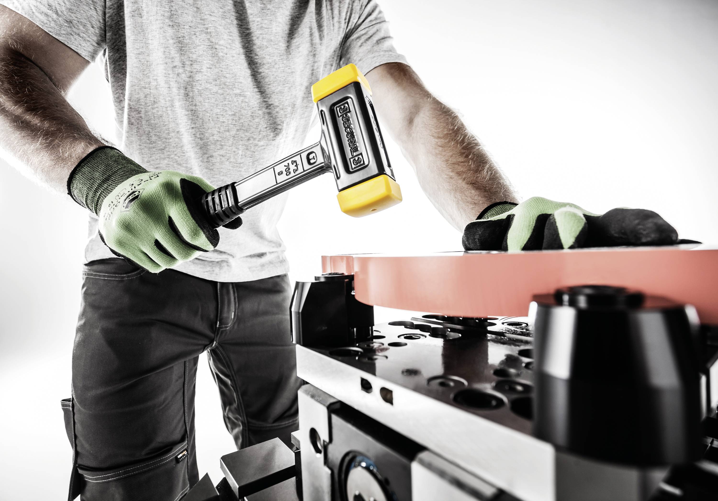 Osoba ubrana w strój roboczy manipuluje żółtym młotkiem przy metalowej szynie na przemysłowym stole.