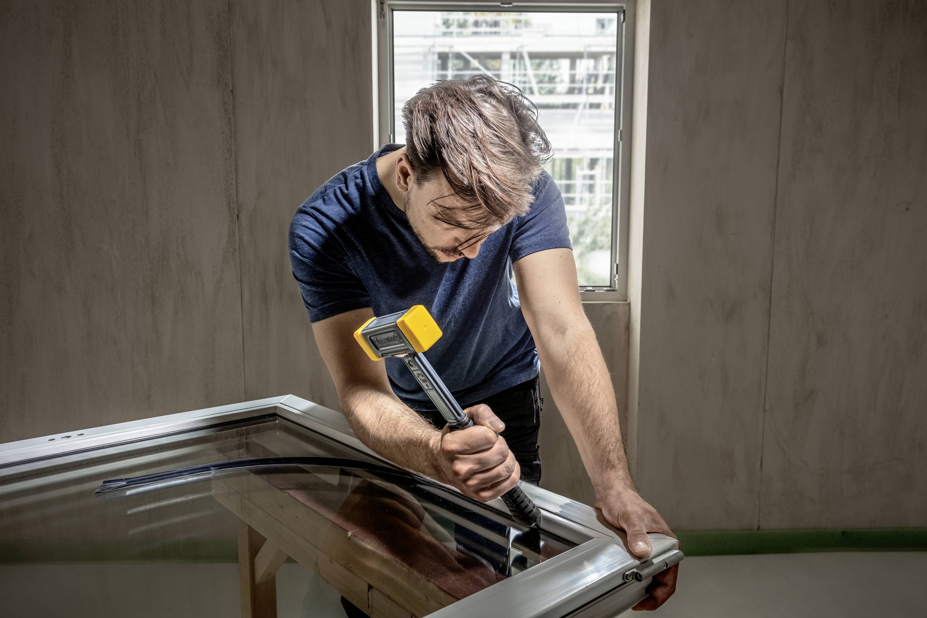 Osoba naprawia ramę okienną, trzyma młotek w ręku i pracuje z dużą koncentracją. W tle połowa okna.