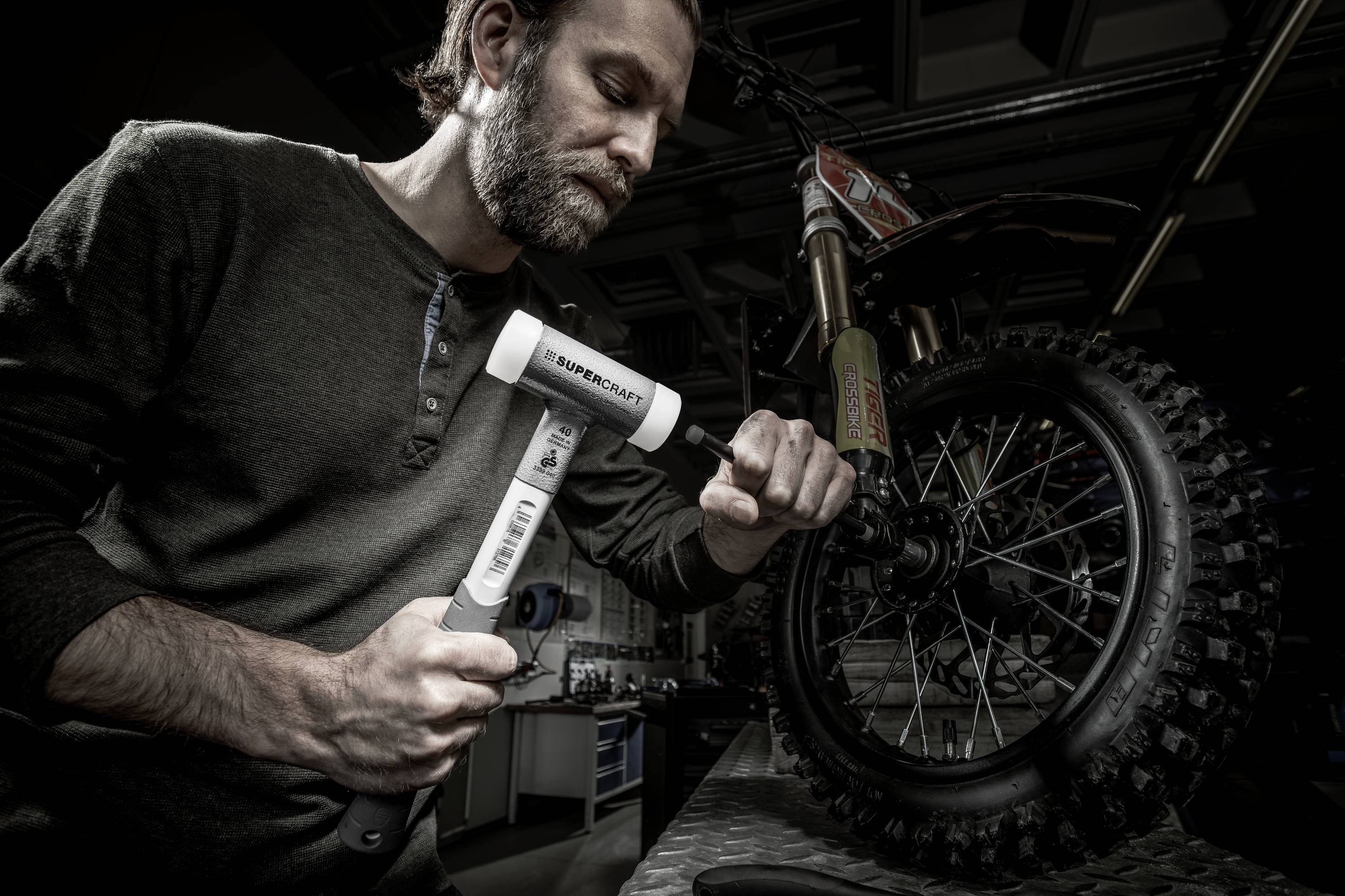 Mężczyzna naprawia motocykl, uderzając białym młotkiem w narzędzie. Motocykl stoi na stole warsztatowym.