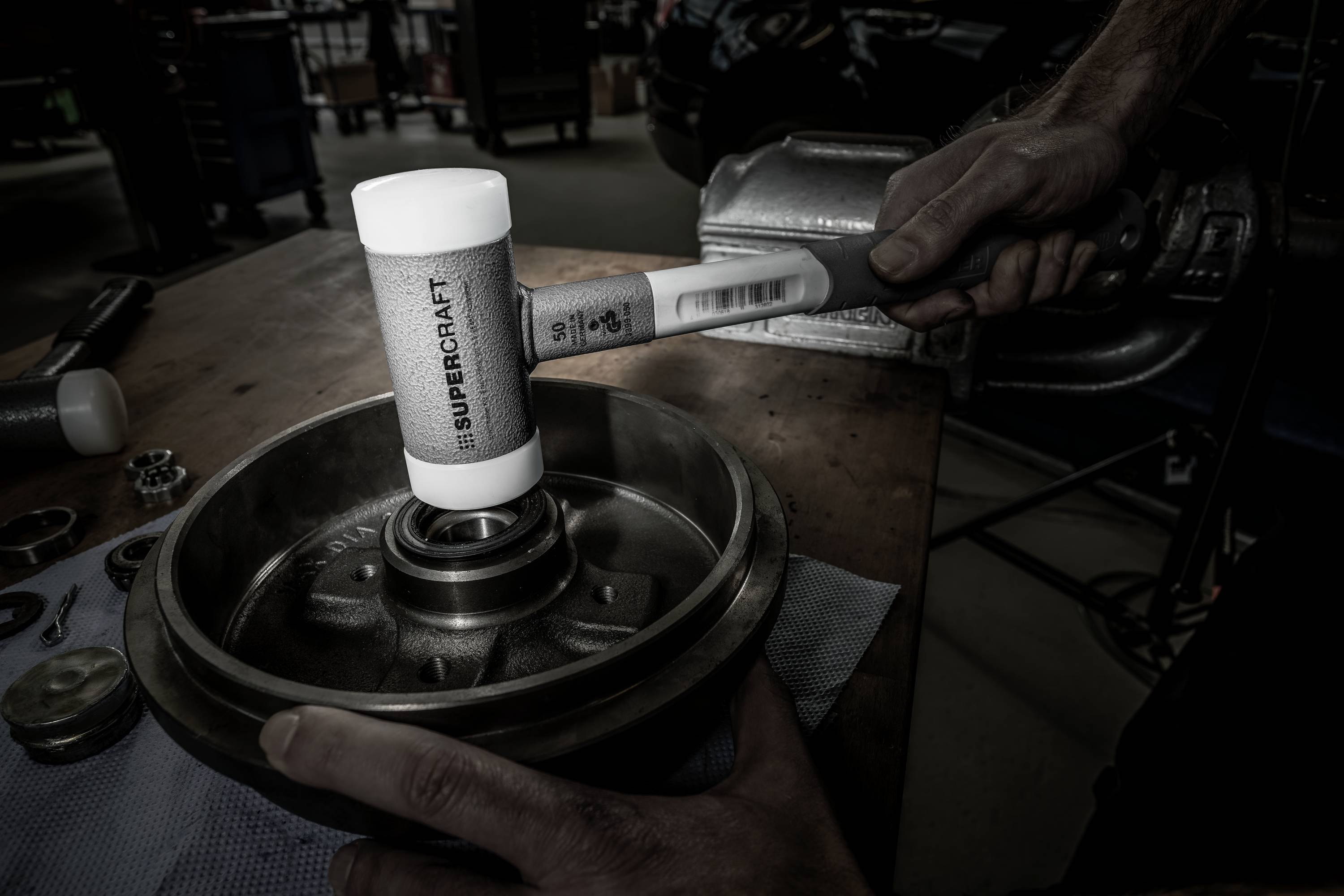 Mechanik obrabia element metalowy na stole warsztatowym białym młotkiem gumowym. W tle widoczne są narzędzia i części samochodowe.