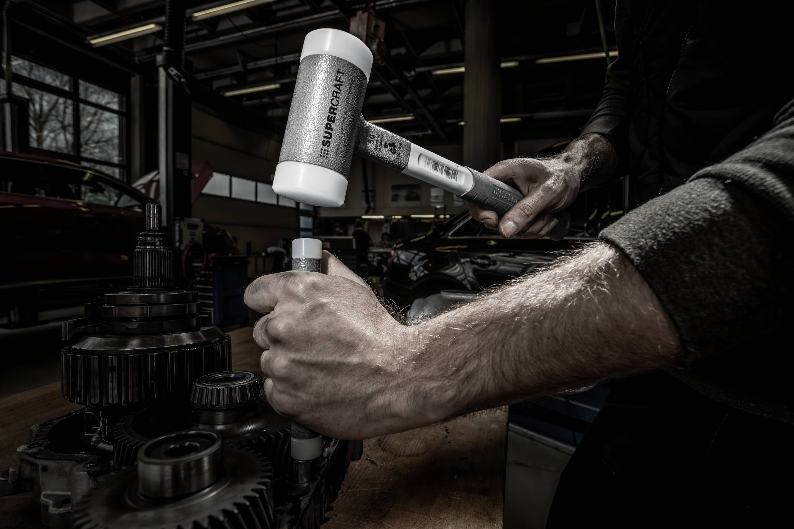 Osoba pracuje białym młotkiem gumowym przy skrzyni biegów w warunkach warsztatowych.