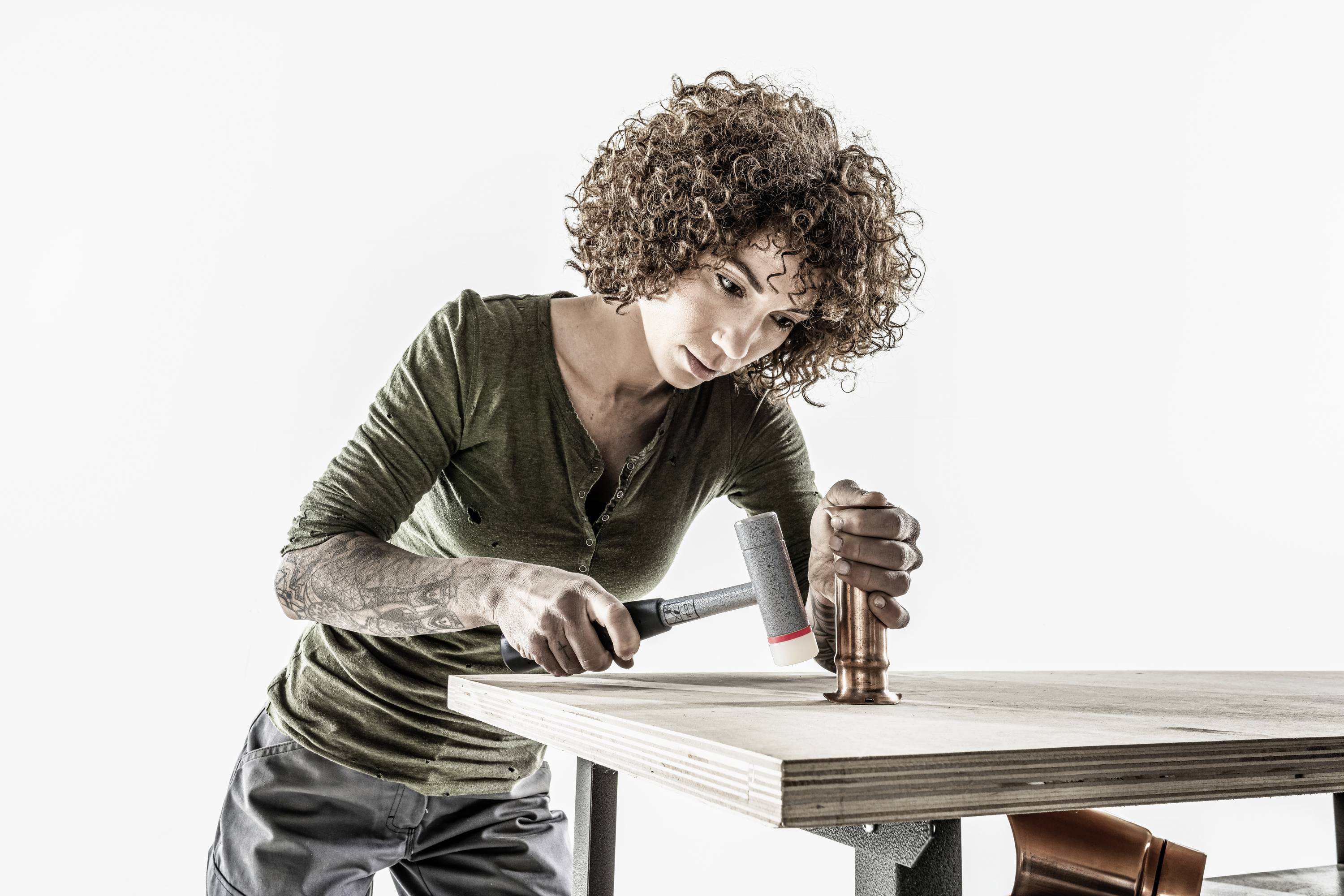 Osoba o kręconych włosach pracuje skupiona, uderzając młotkiem w miedzianą rurę leżącą na drewnianym stole.