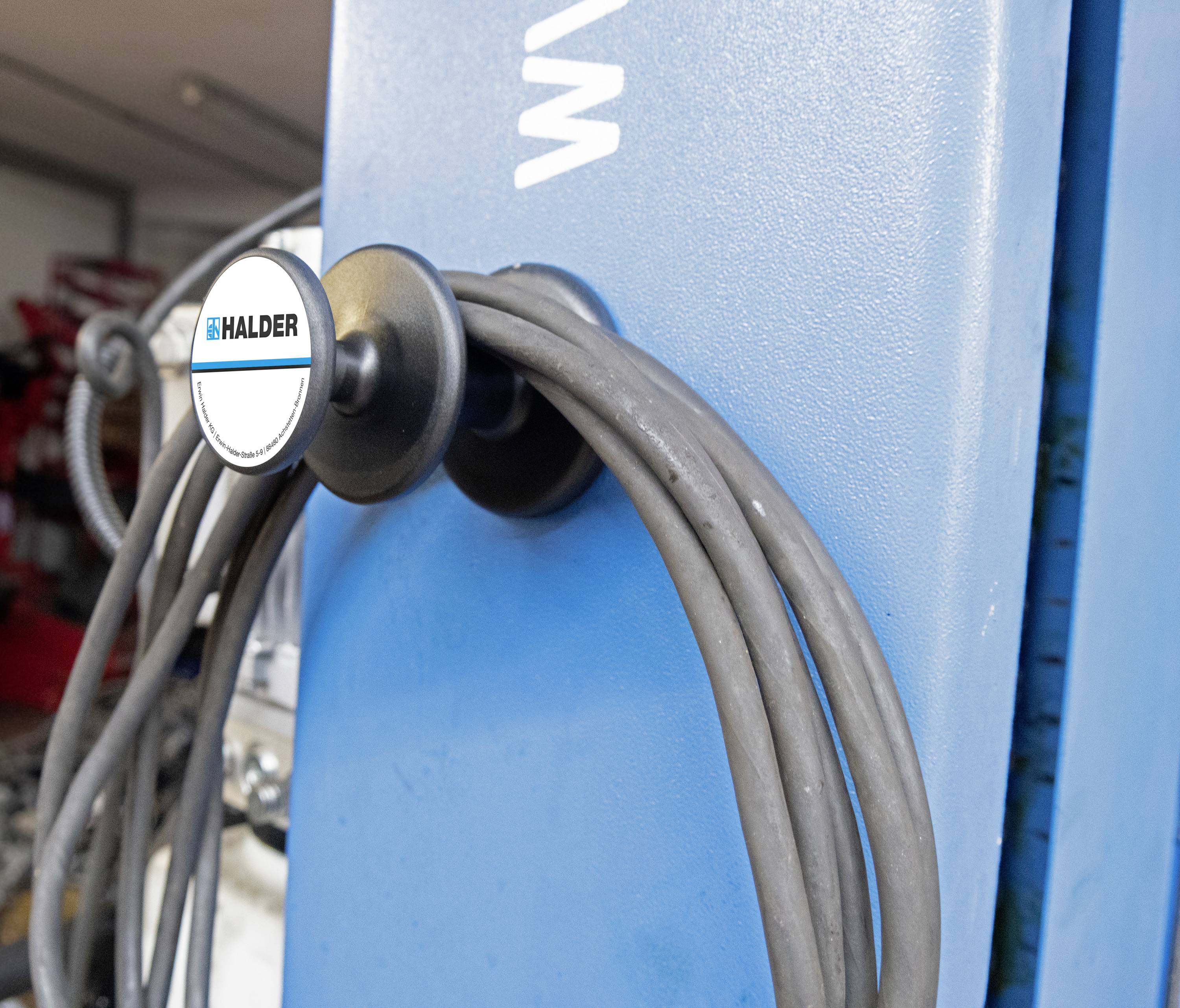 Szary kabel z etykietą Haldera przymocowany do niebieskiego urządzenia, prawdopodobnie w warsztacie lub środowisku przemysłowym.