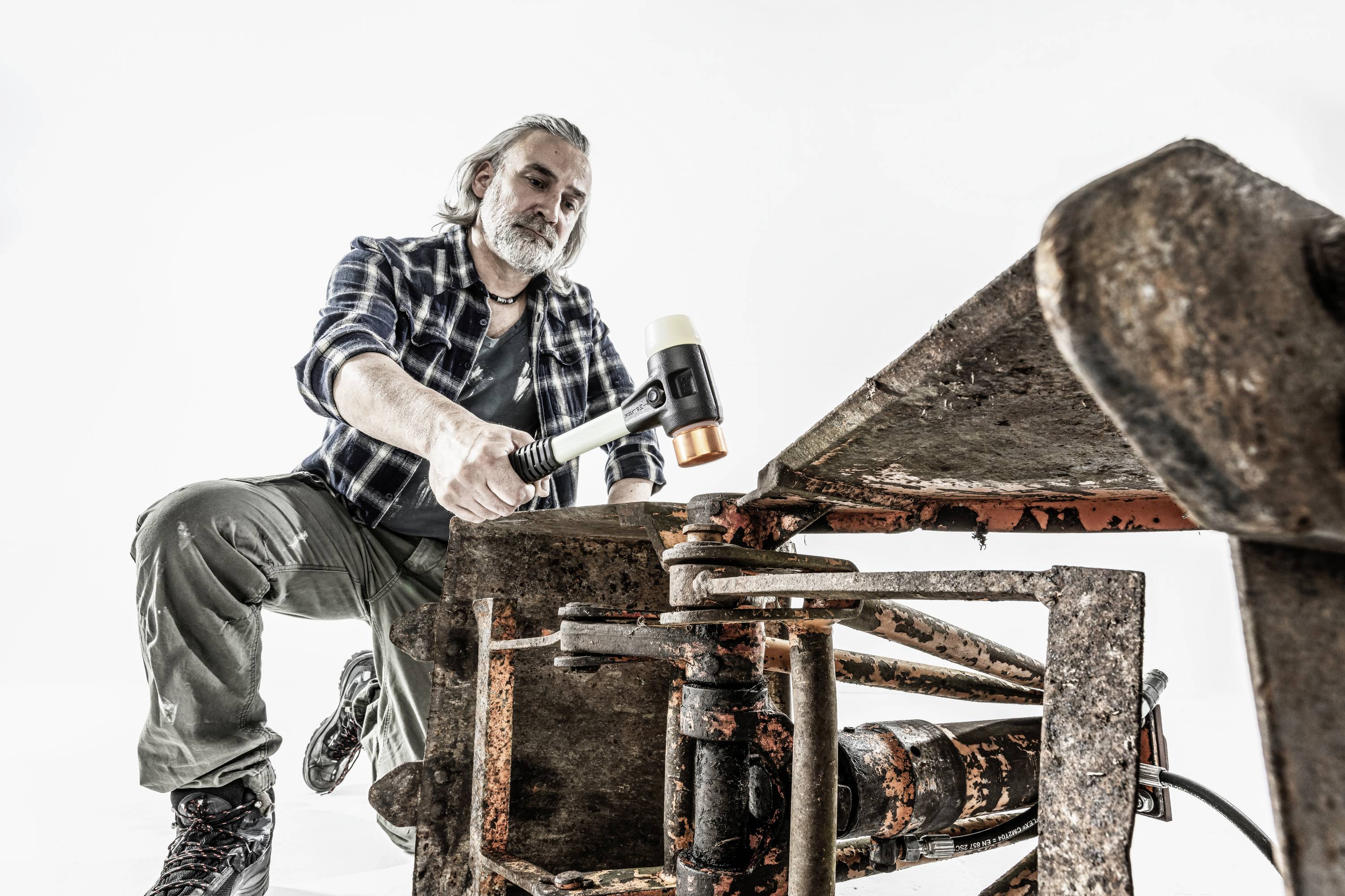 Mężczyzna z siwymi włosami w warsztacie obija młotkiem dużą metalową część. Ma na sobie koszulę w kratkę.