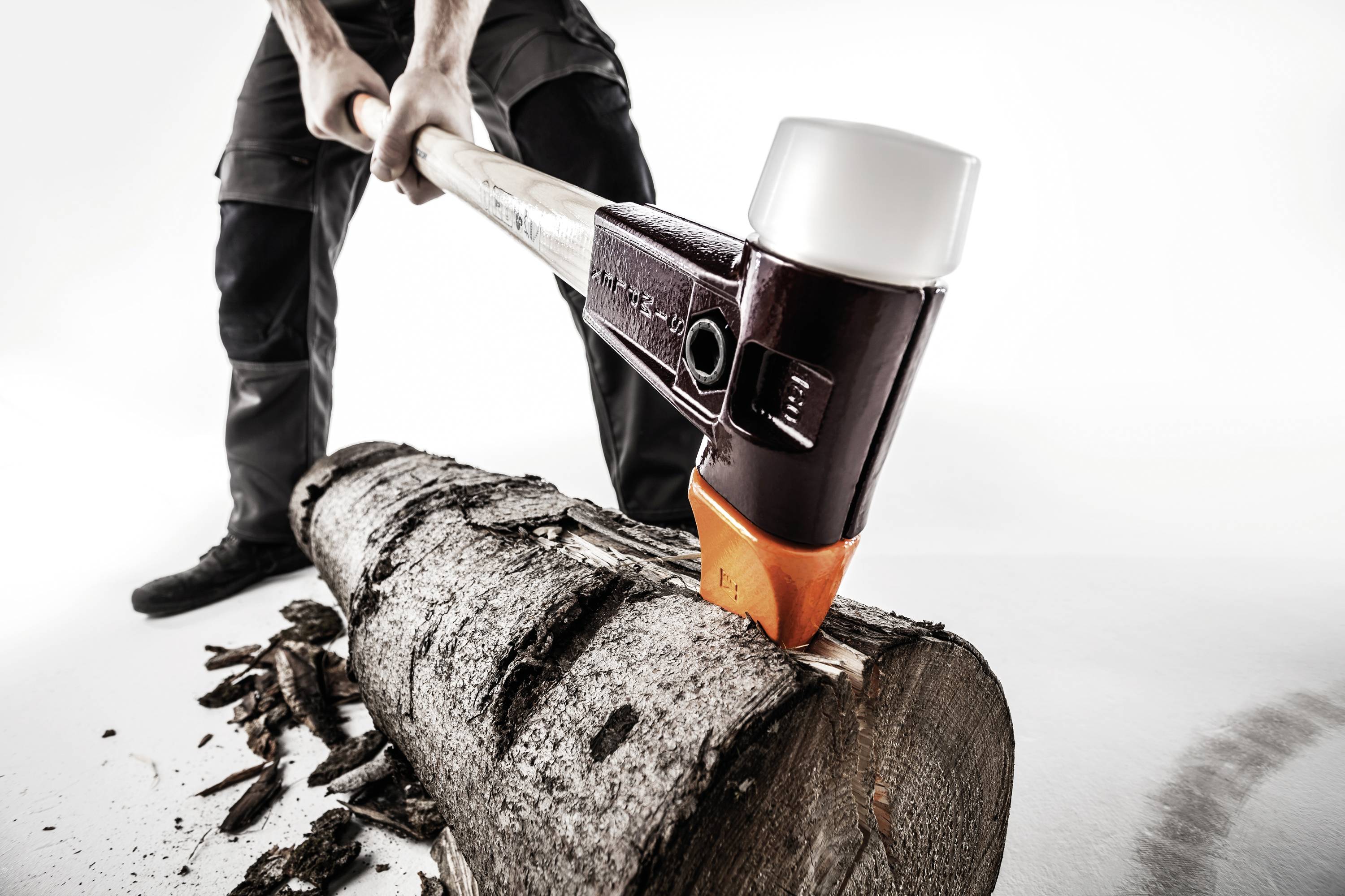 Osoba rozłupuje grubą kłodę drewna siekierą. Na ziemi leżą wióry drewniane.