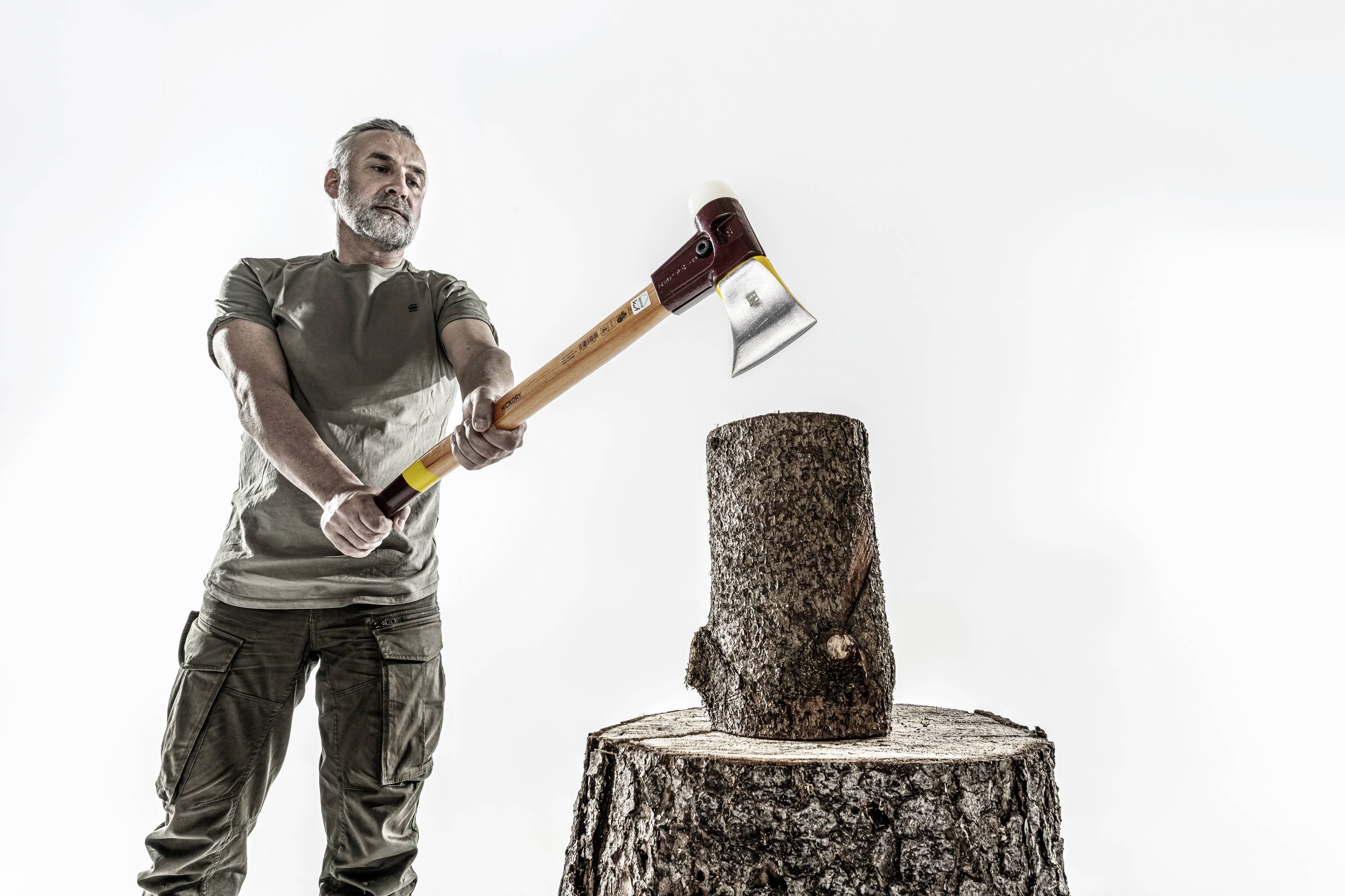 Mężczyzna w koszulce i spodniach podnosi siekierę, aby rozłupać drewniany kloc na pniu drzewa.