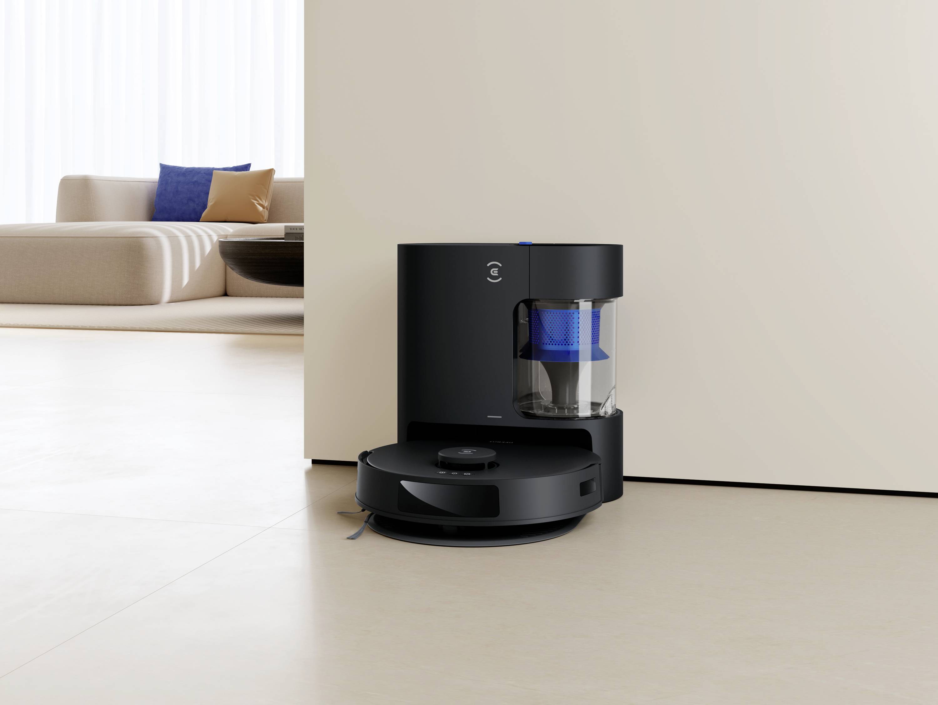 Czarny robot sprzątający z bazą ładującą stoi w nowoczesnym, minimalistycznie urządzonym salonie z beżową sofą i niebieskimi poduszkami.