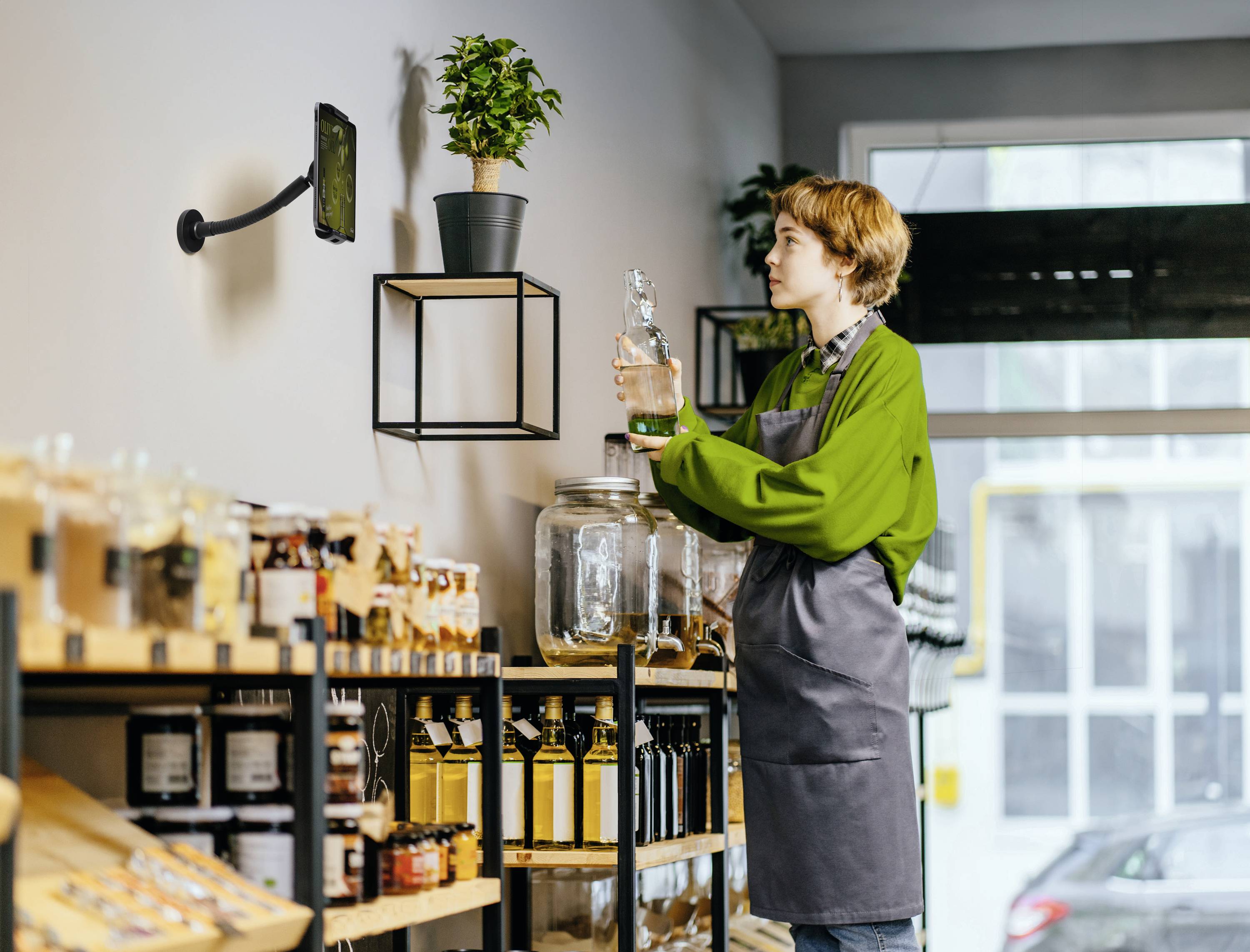Osoba w zielonej koszuli i fartuchu stoi w sklepie ekologicznym przy regale, trzymając w ręku wielorazową szklną butelkę, aby napełnić ją produktami.