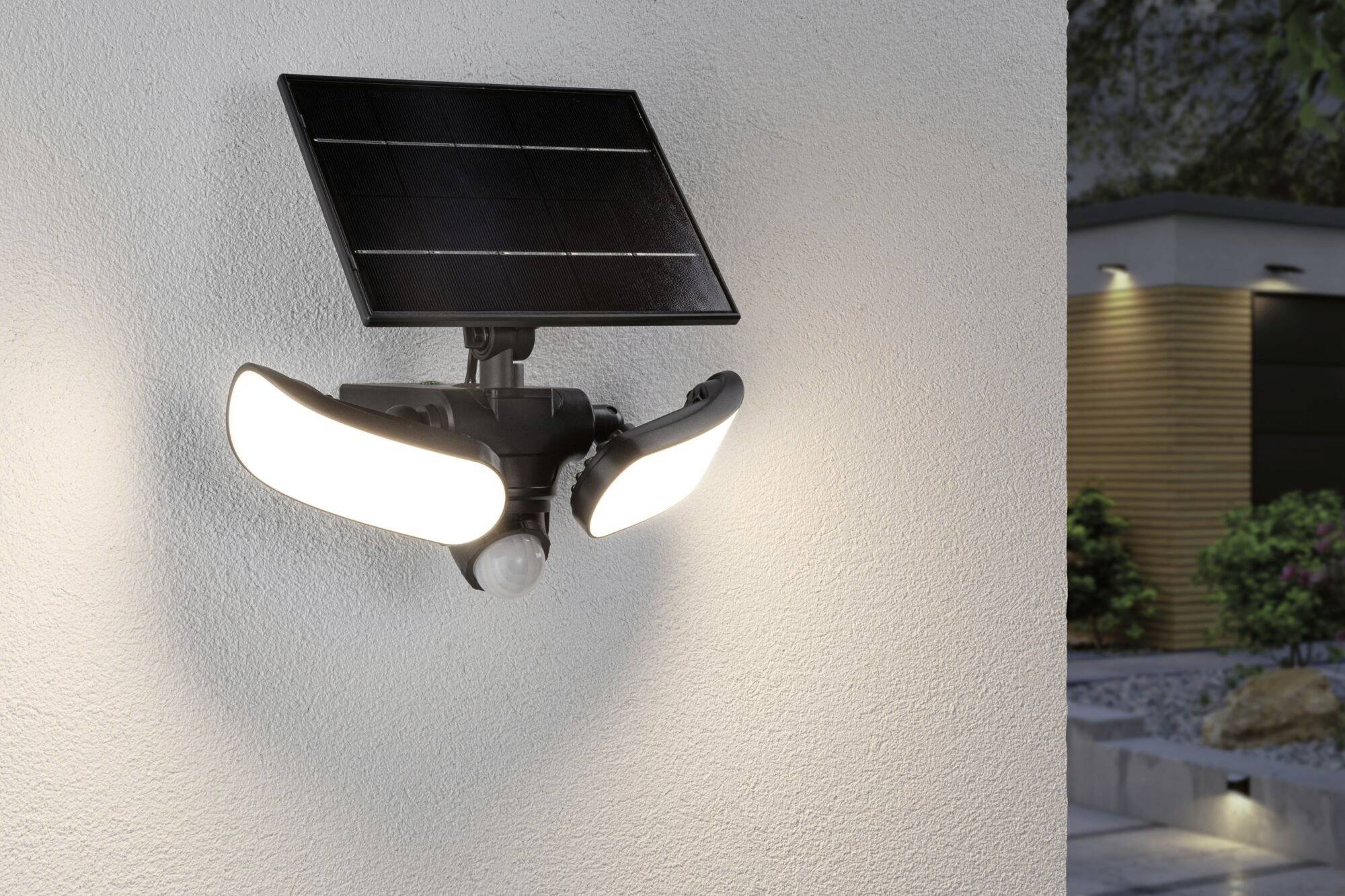 Lampa solarna z dwoma reflektorami i czujnikiem ruchu zamontowana na ścianie domu, oświetlająca fragment nowoczesnego ogrodu.