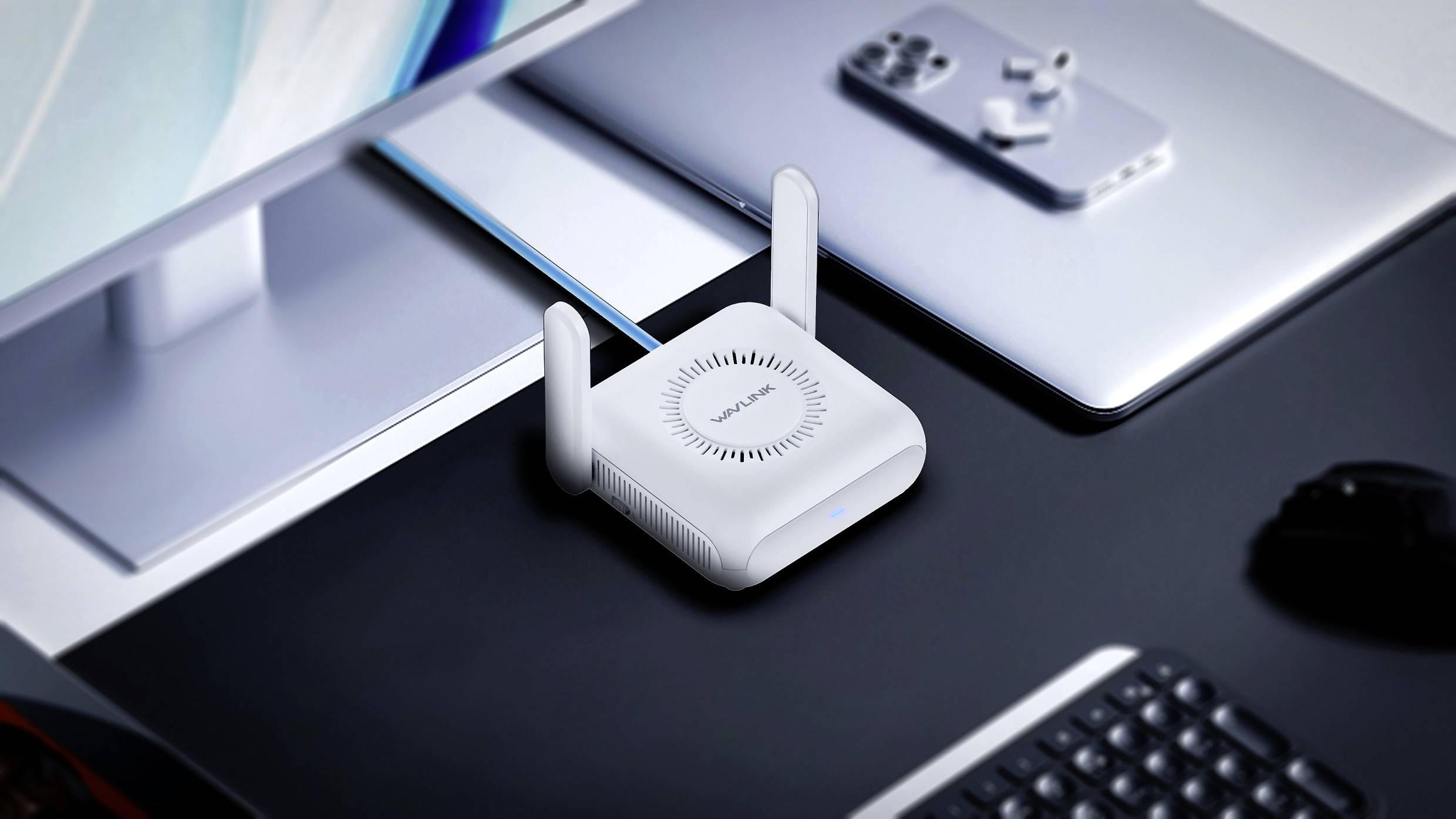 Biały router WLAN z dwoma antenami stoi na biurku obok laptopa, klawiatury i monitora.