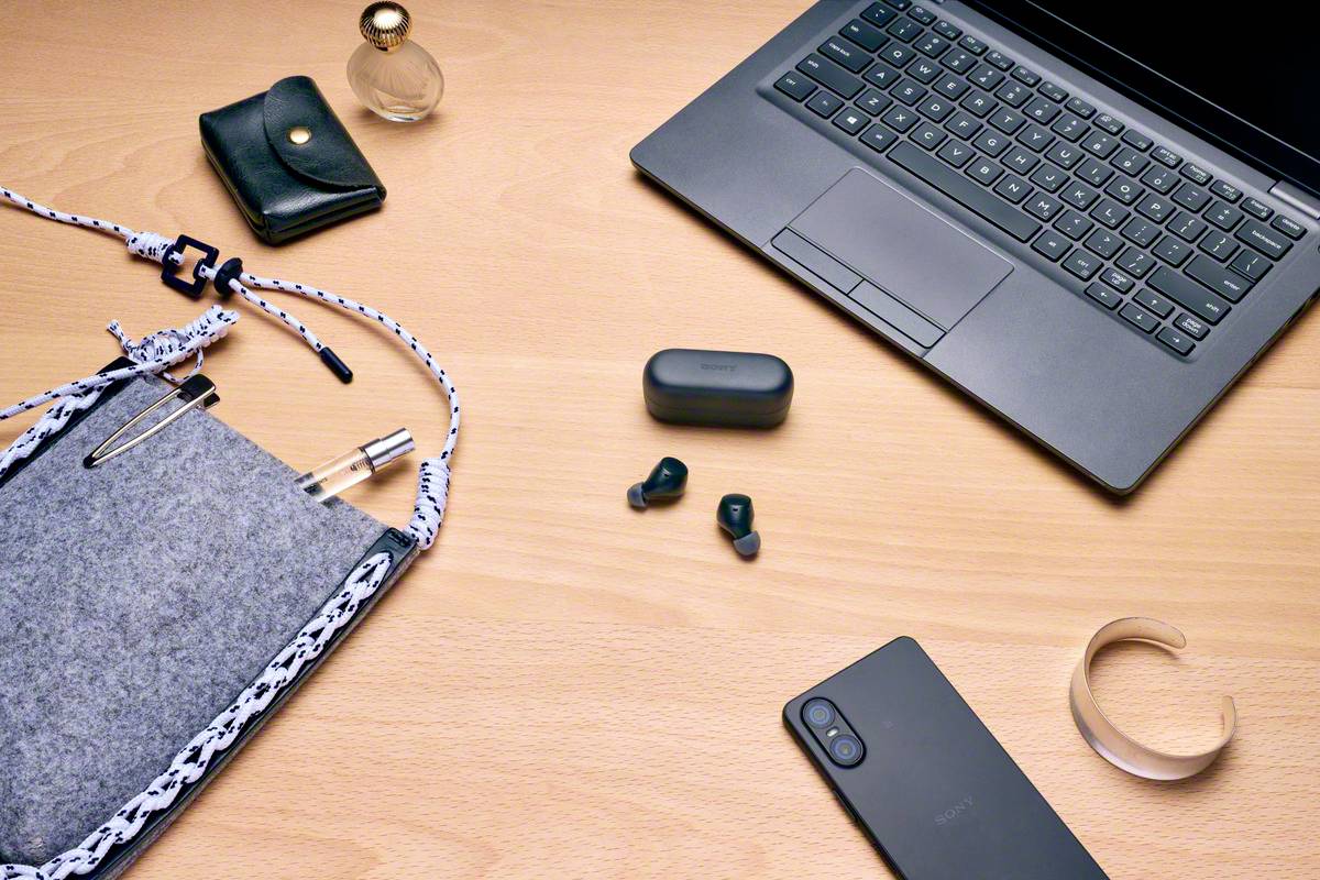 Biurko z laptopem, smartfonem, słuchawkami, notatnikiem, flakonem perfum i portfelem. Nacisk na nowoczesną technologię i akcesoria.