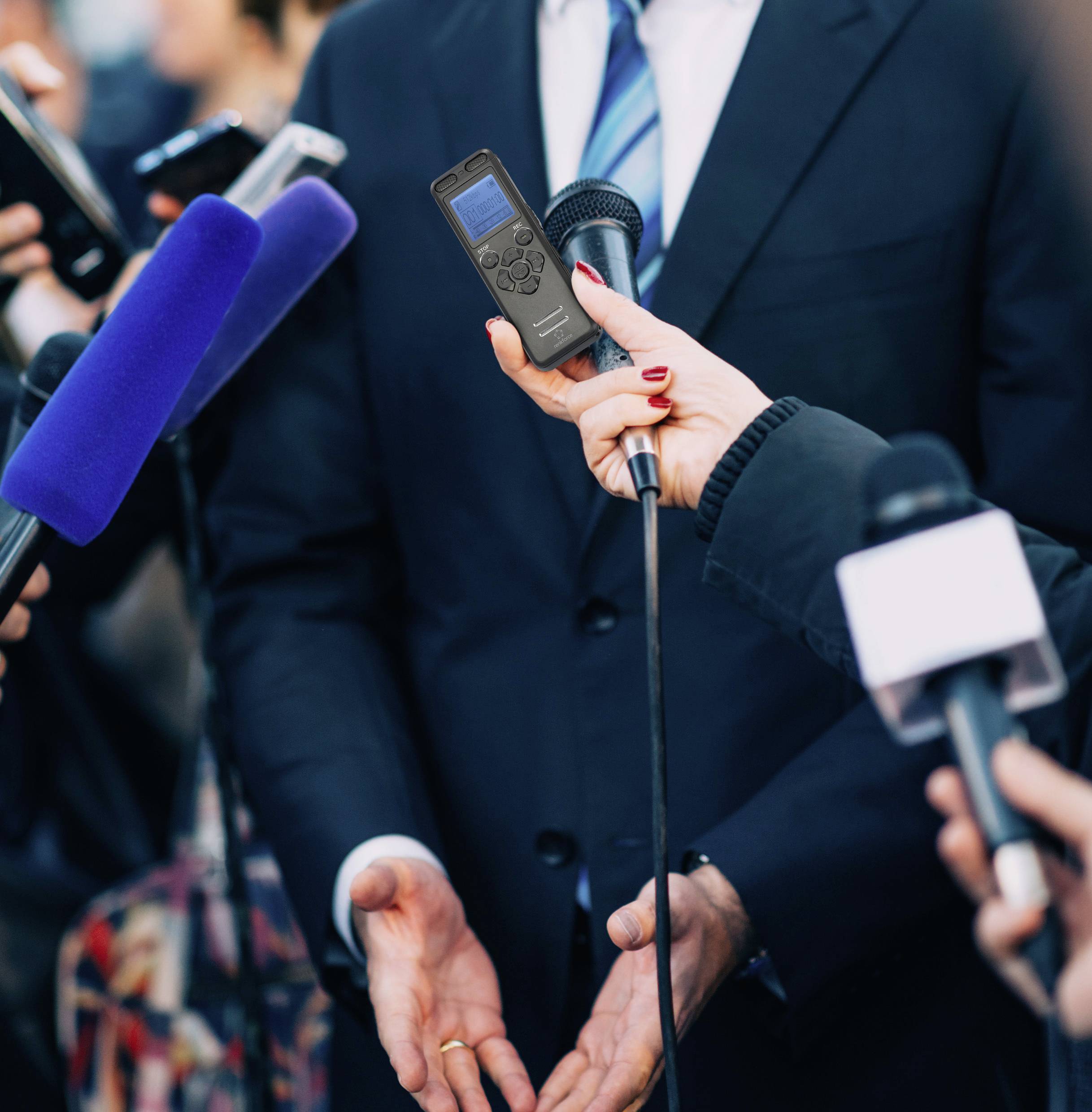 Osoba w garniturze udziela wywiadu przed reporterami, którzy kierują w jej stronę mikrofony.