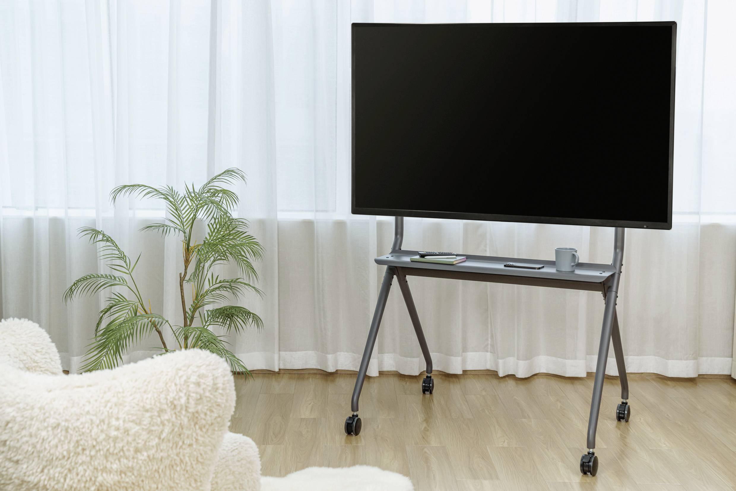 Płaski telewizor na jezdnym stojaku w jasnym pomieszczeniu z drewnianą podłogą. Na tle widać fotel i roślinę.