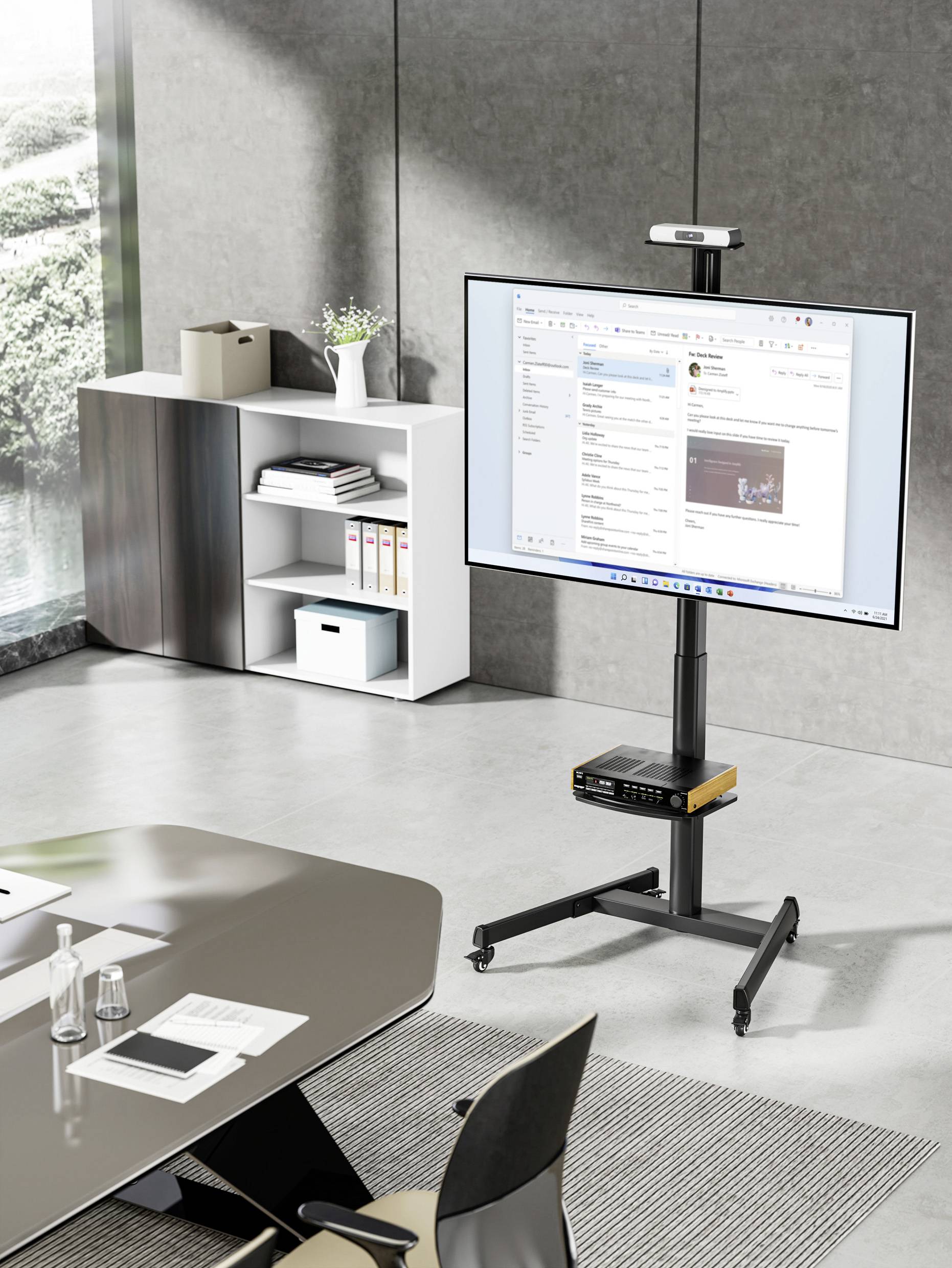 Sala konferencyjna z dużym ekranem na stojaku, wyświetlającym aplikację pocztową. W tle szafa z półkami.