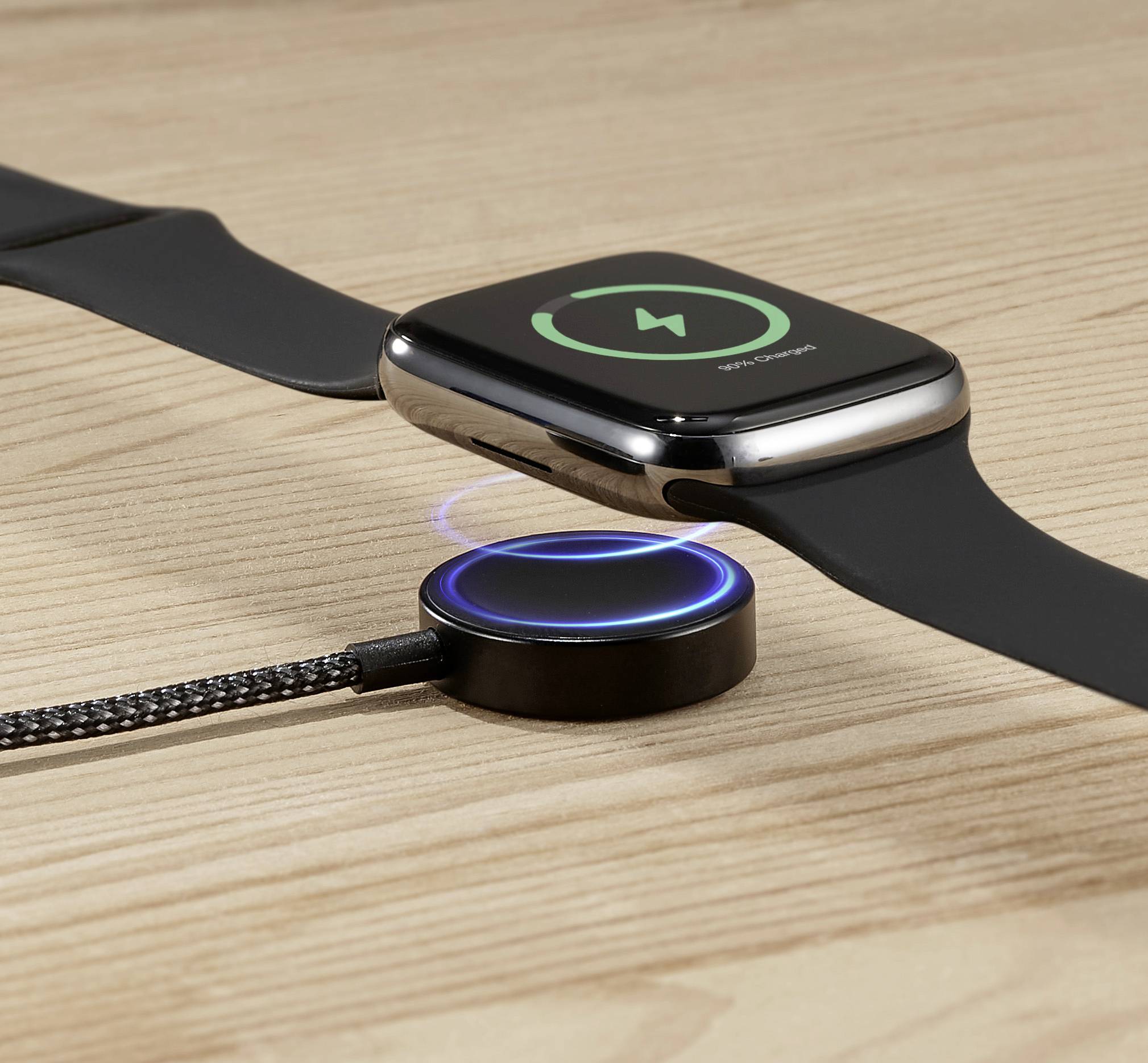 Smartwatch leży na drewnianym stole i jest ładowana bezprzewodowo. Ładowarka z podświetlonym pierścieniem wskazuje postęp ładowania.