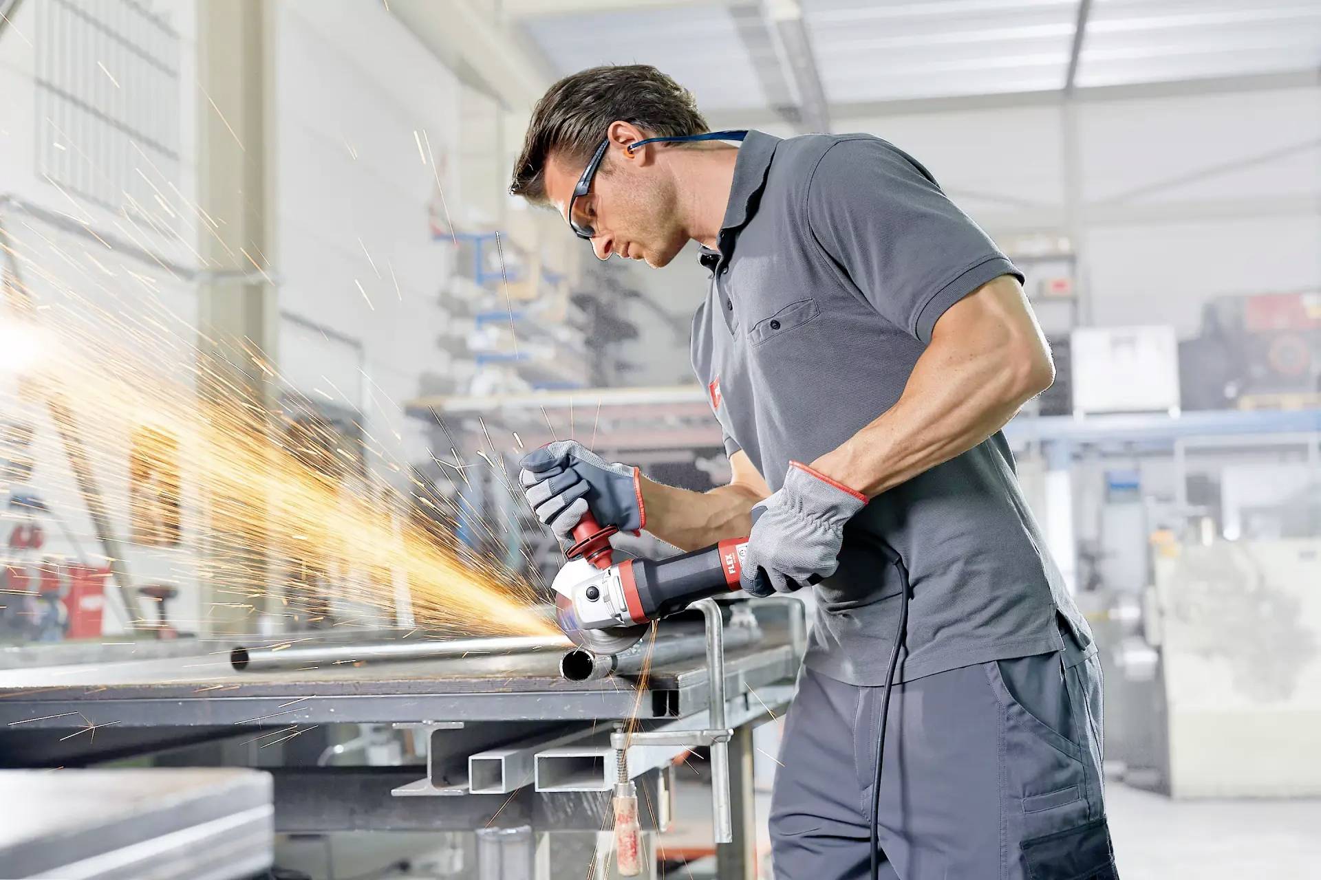 Mężczyzna w okularach ochronnych szlifuje kawałek metalu szlifierką kątową w warsztacie, powodując powstawanie iskier.