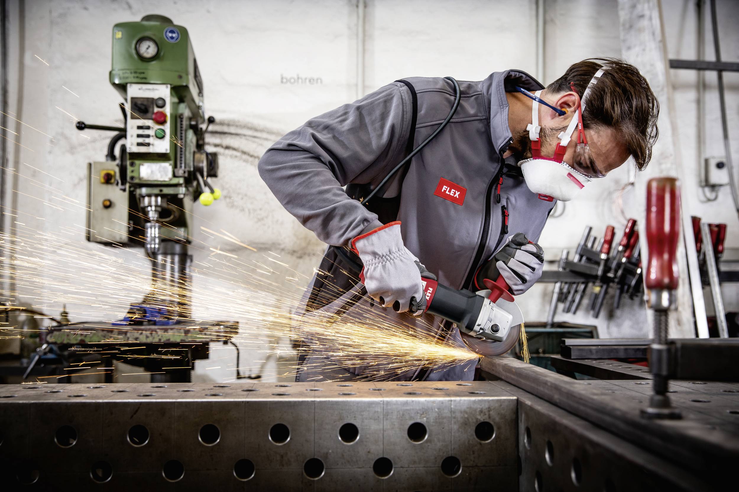 Mężczyzna pracuje z szlifierką kątową na płycie metalowej. Lecą iskry. Nosi odzież ochronną i maskę przeciwpyłową.