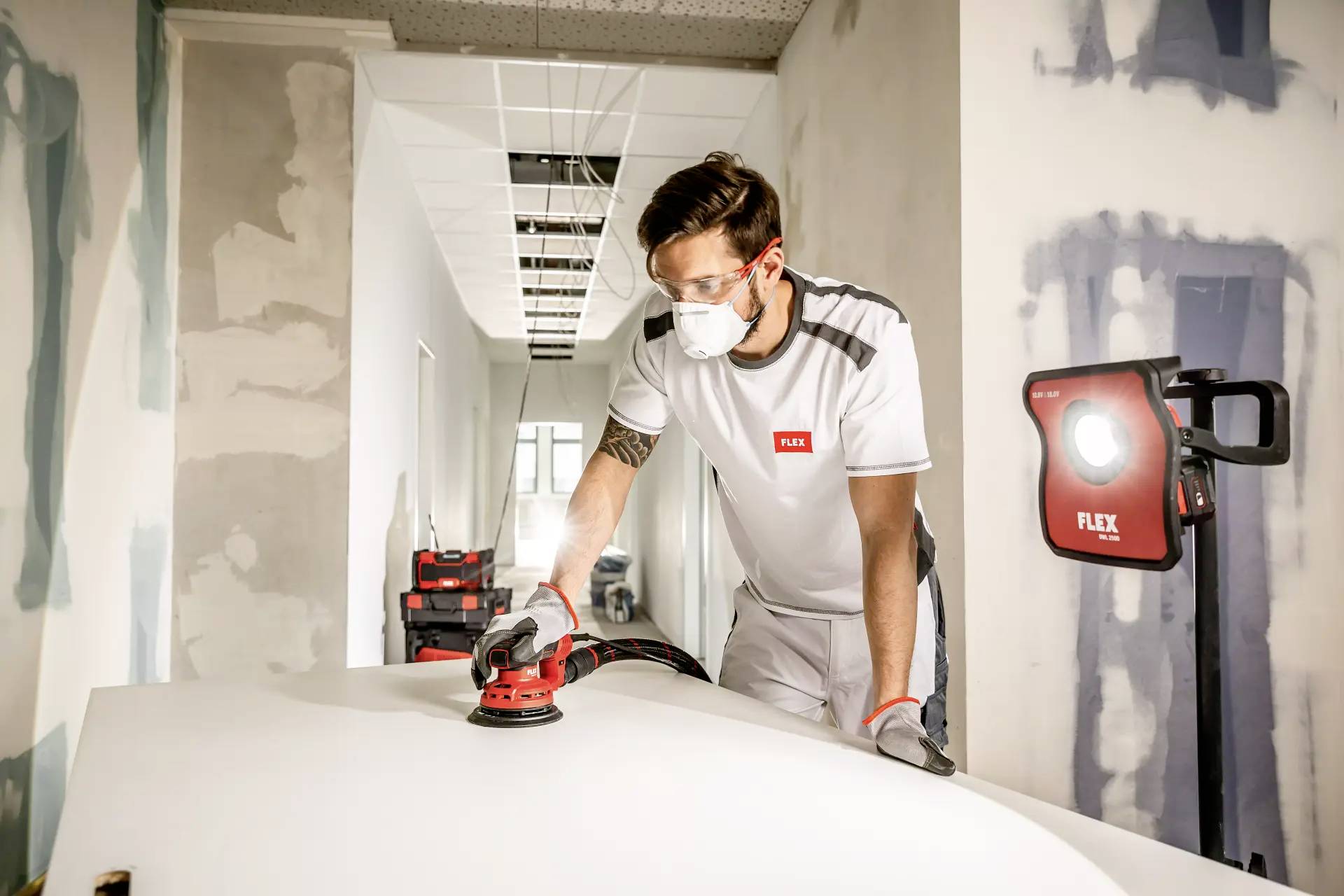 Mężczyzna w ubraniu roboczym szlifuje białą powierzchnię elektronarzędziem w częściowo wyremontowanym pomieszczeniu. Nosi okulary ochronne i maskę.