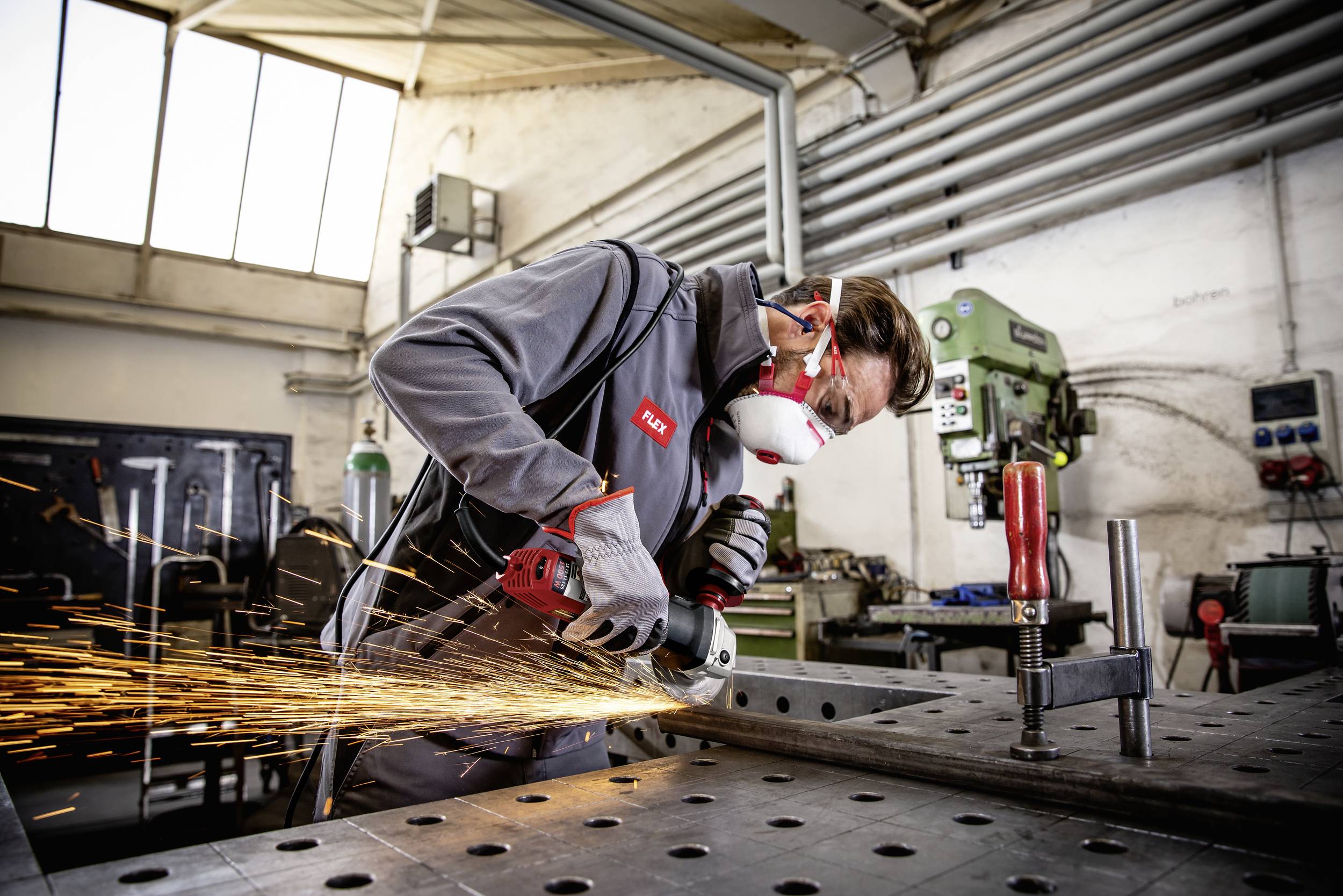 Robotnik w odzieży ochronnej szlifuje metal na warsztacie przy stole warsztatowym. Wokół niego lecą iskry, w tle widać maszyny.