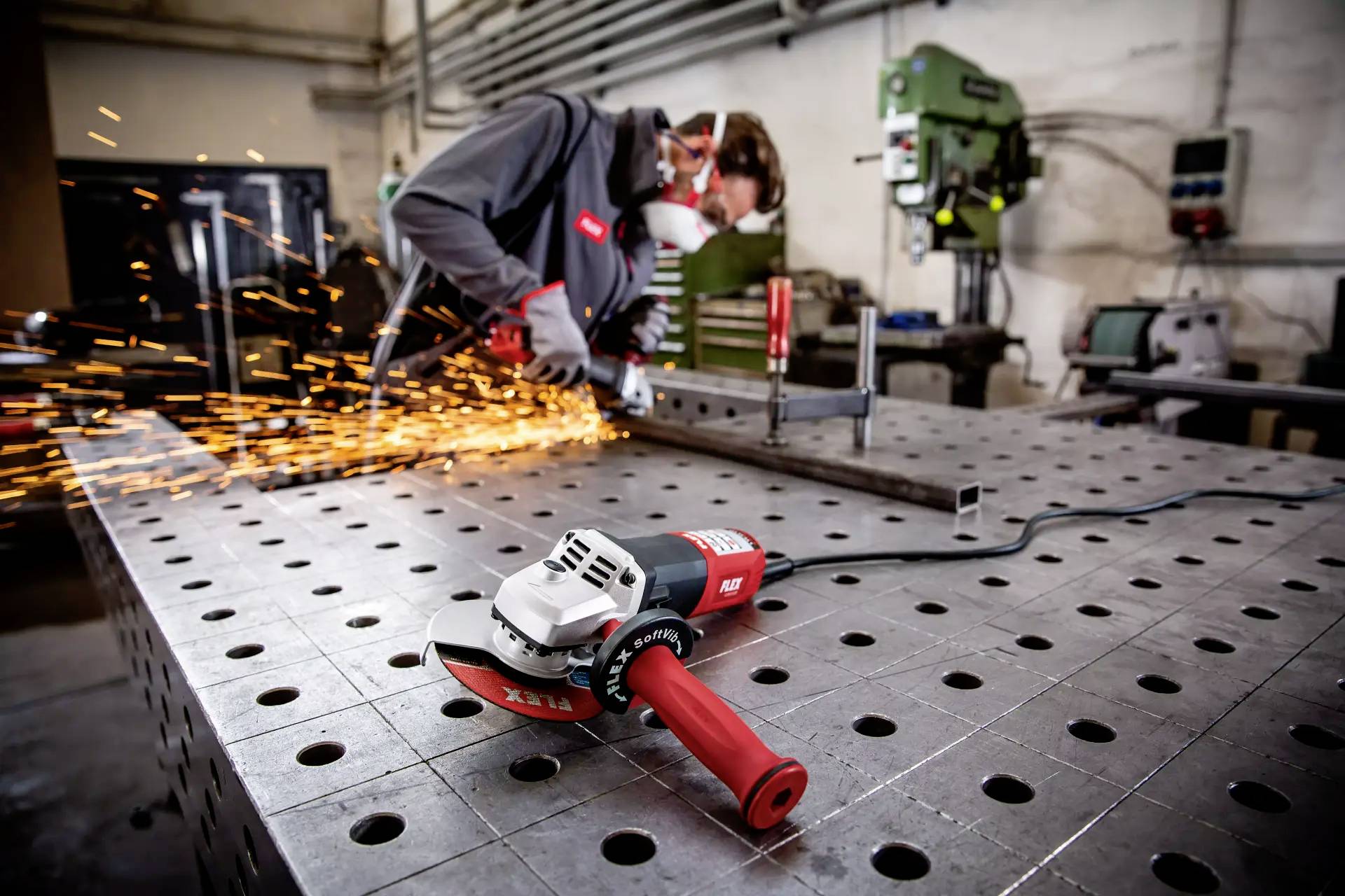 Robotnik spawający metal w warsztacie, iskry się sypią. Na pierwszym planie leży nieużywana szlifierka kątowa na metalowym stole.