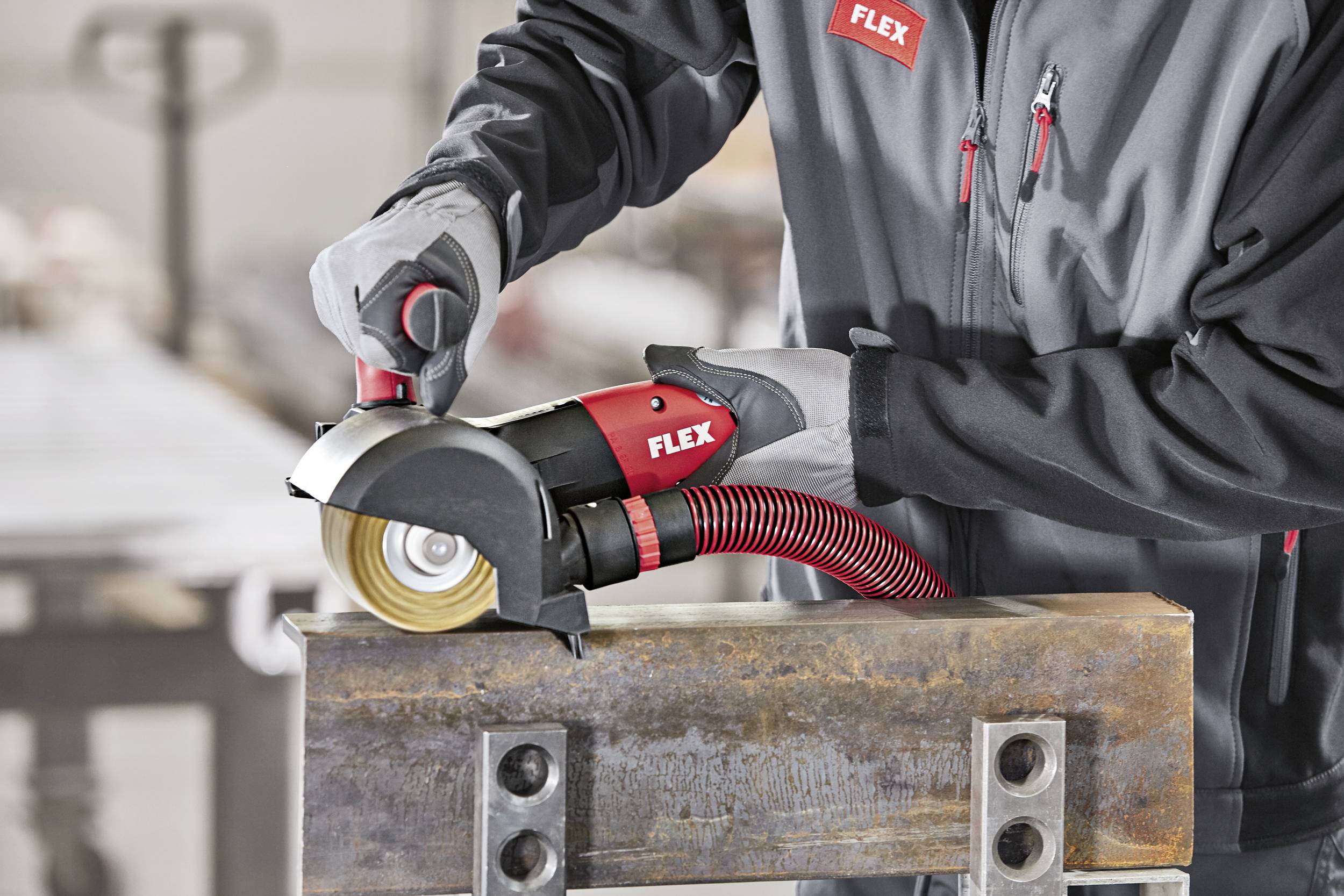 Osoba pracuje z szlifierką kątową FLEX na metalowej konstrukcji. Urządzenie jest koloru czerwonego i czarnego, a pracownik jest ubrany w odzież ochronną.