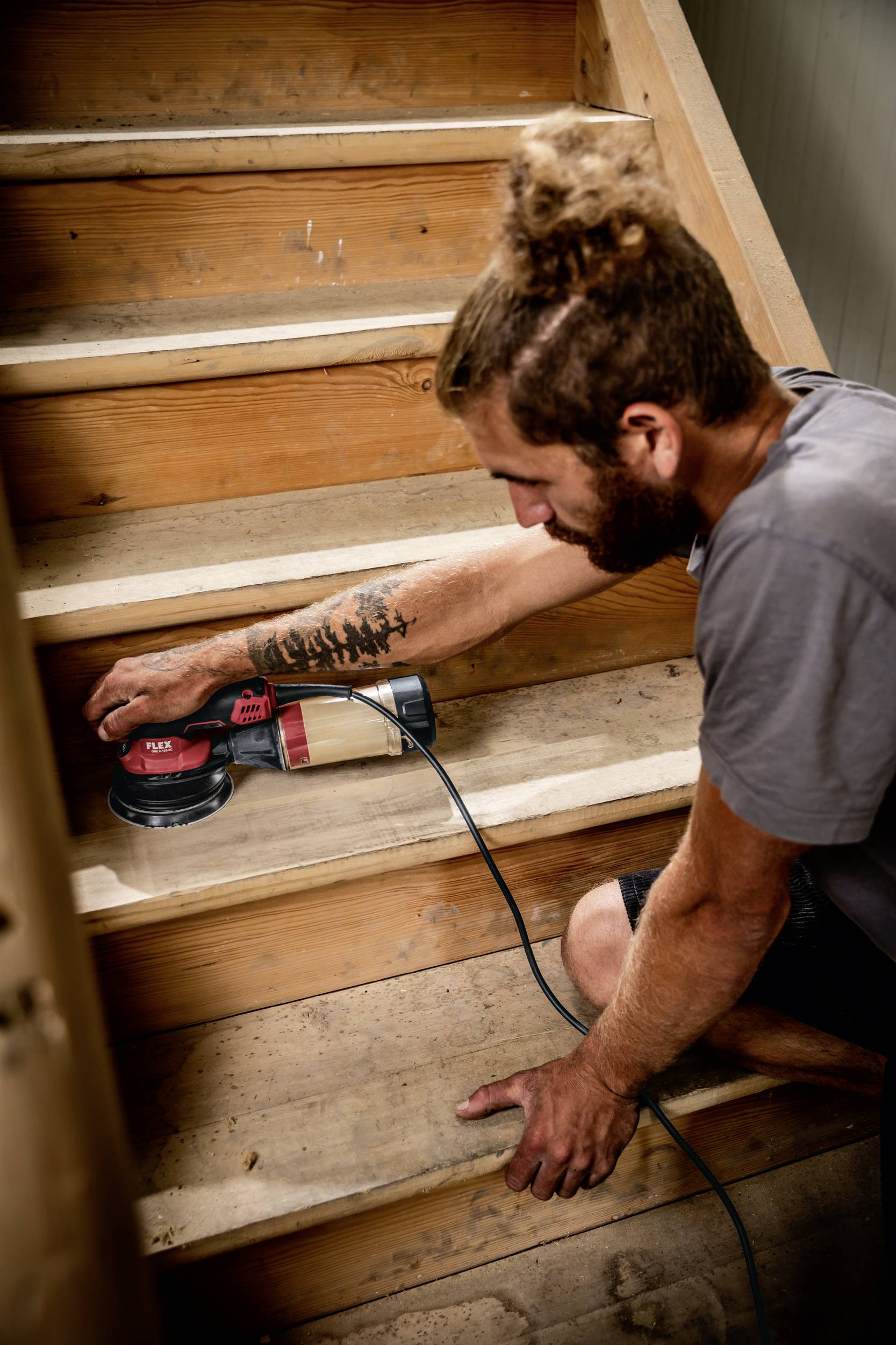 Osoba szlifuje drewniane schody za pomocą elektrycznej szlifierki. Ma tatuaż na ramieniu i nosi szary t-shirt.