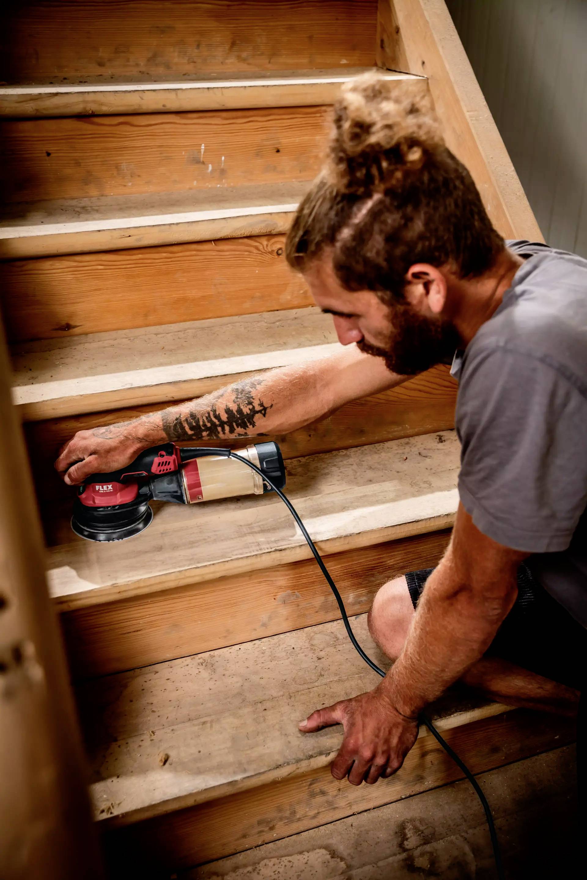 Mężczyzna szlifuje drewniane schody elektryczną szlifierką. Ma na sobie swobodny strój i jest całkowicie skupiony na wykonywanej pracy.