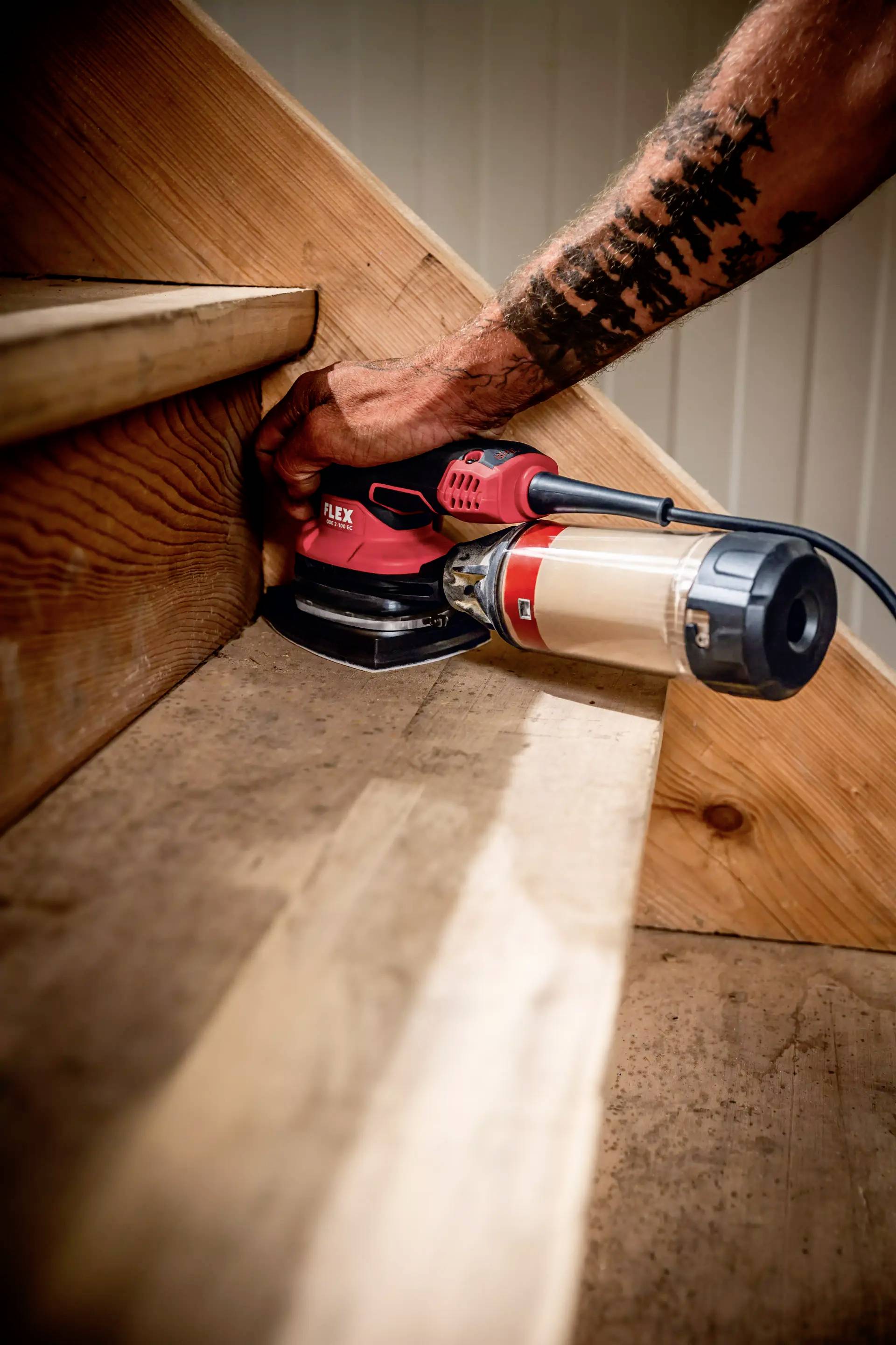 Osoba z tatuażami wygładza drewniane stopnie schodów elektryczną szlifierką w odnowionym budynku.