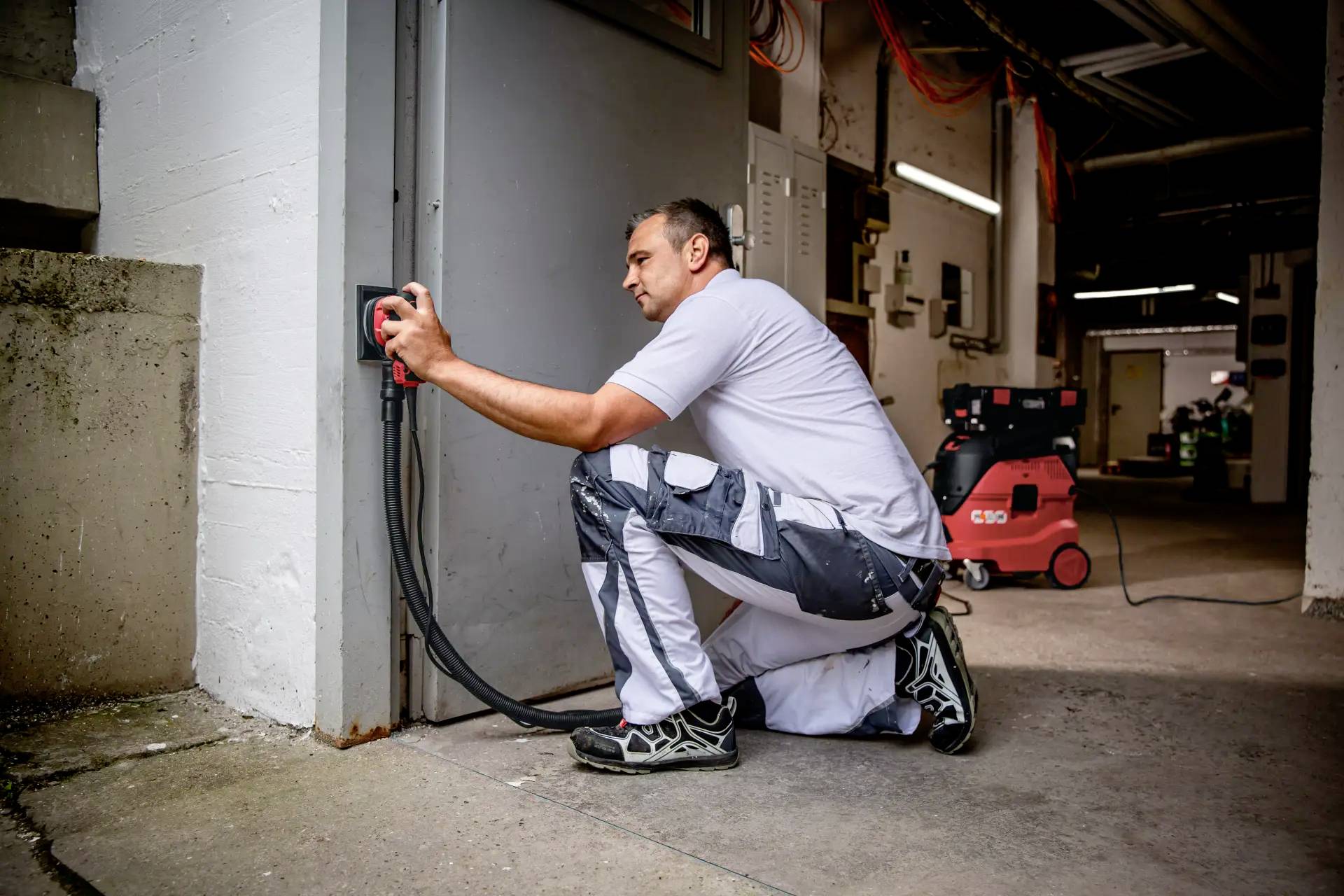 Osoba w stroju roboczym czyści futrynę odkurzaczem w kolorach czerwono-czarnych w środowisku przemysłowym.