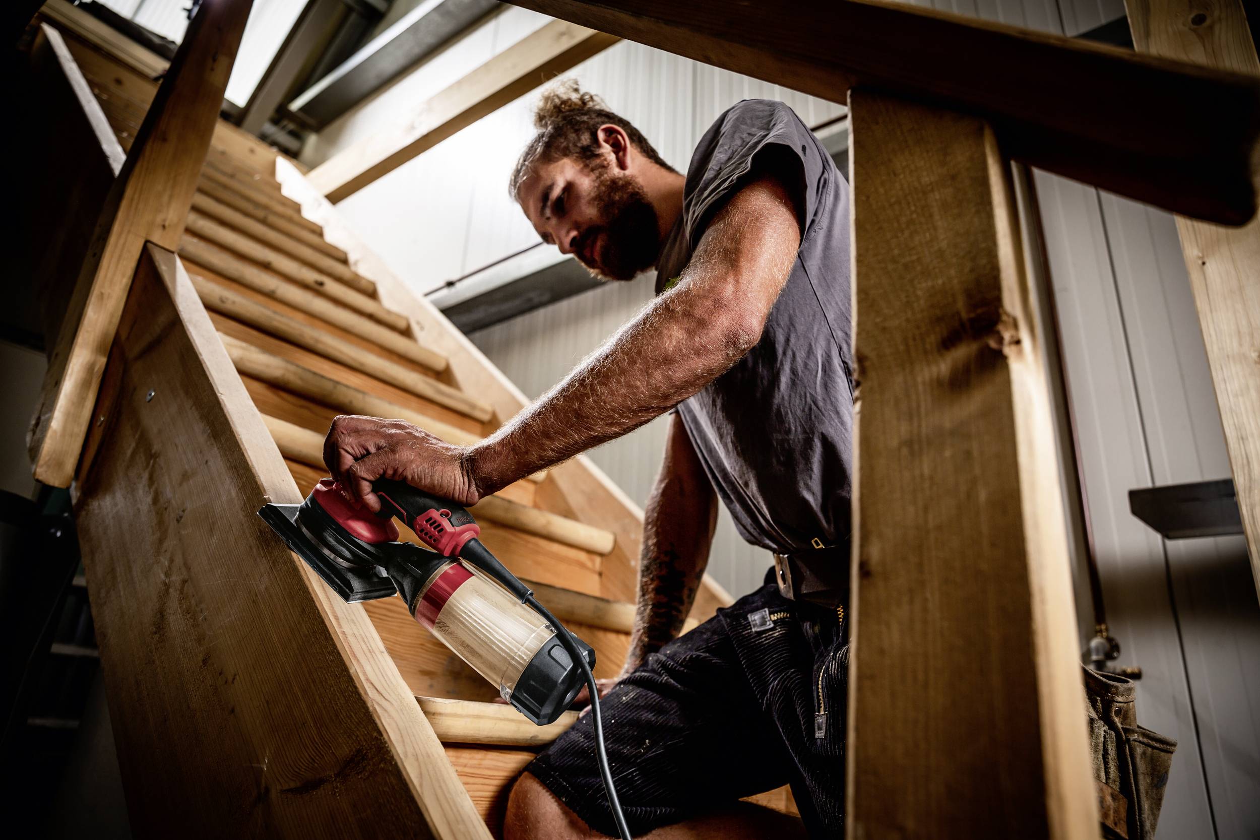 Mężczyzna szlifuje drewnianą poręcz schodów w warsztacie. Ma na sobie szary t-shirt i używa elektrycznego urządzenia szlifierskiego.