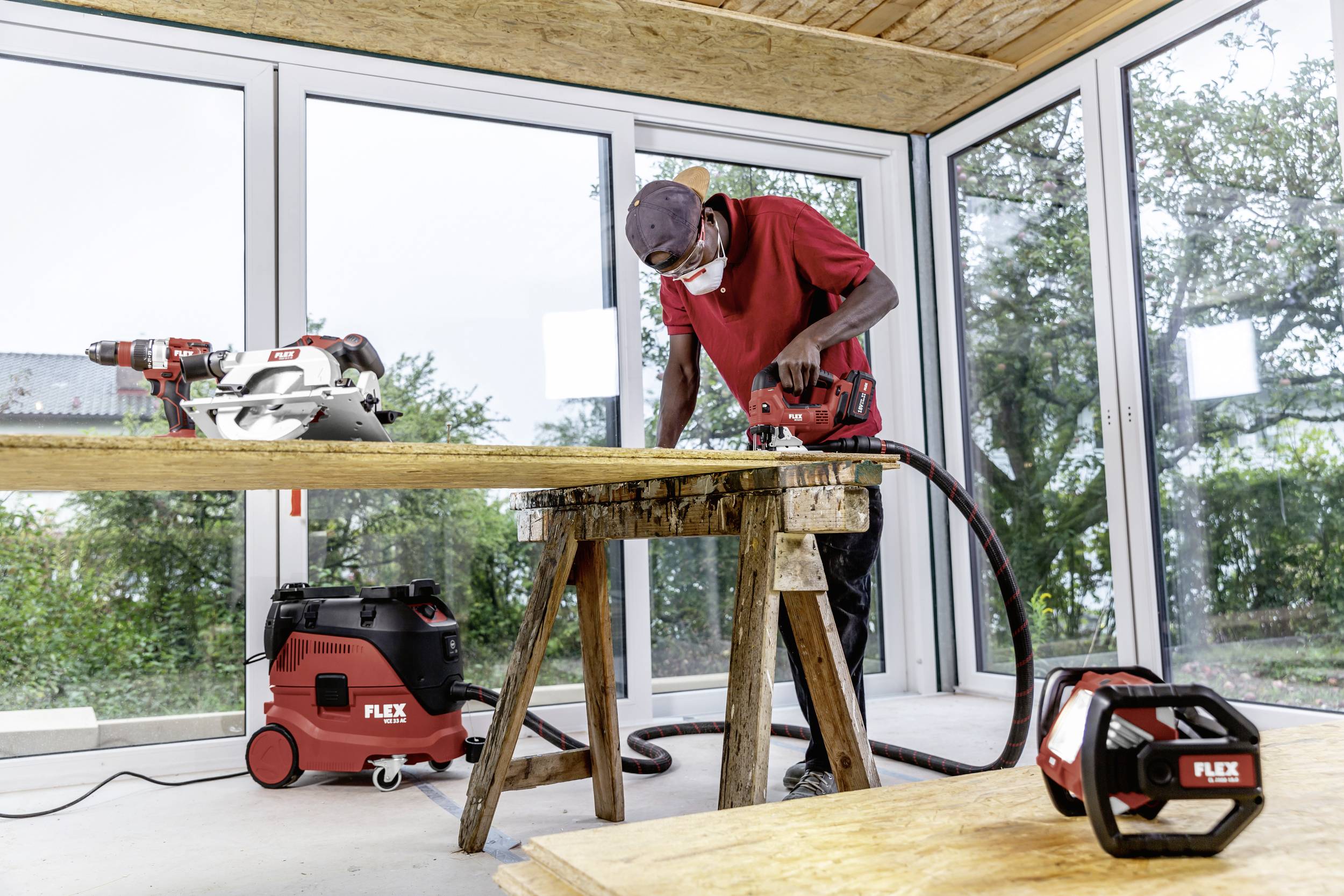 Rzemieślnik tnie drewno elektryczną piłą w jasnym pomieszczeniu z dużymi oknami. Obok niego stoi odkurzacz przemysłowy.