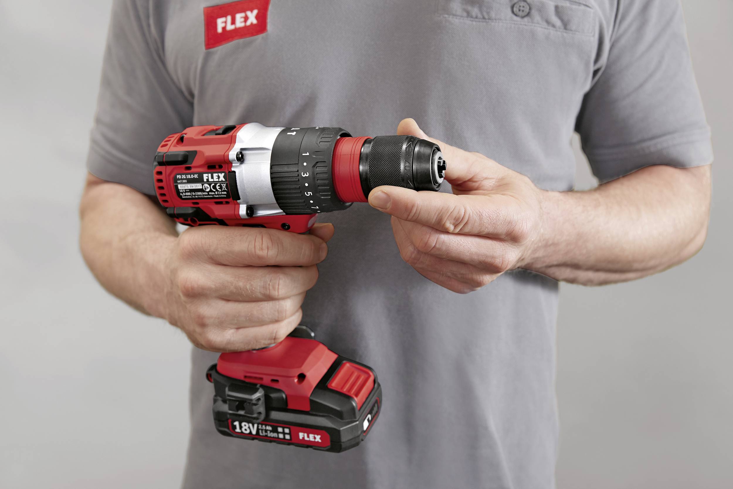 Osoba trzyma czerwony akumulatorowy wkrętak marki Flex. Wkrętak posiada czarne i srebrne akcenty oraz napis '18V'.