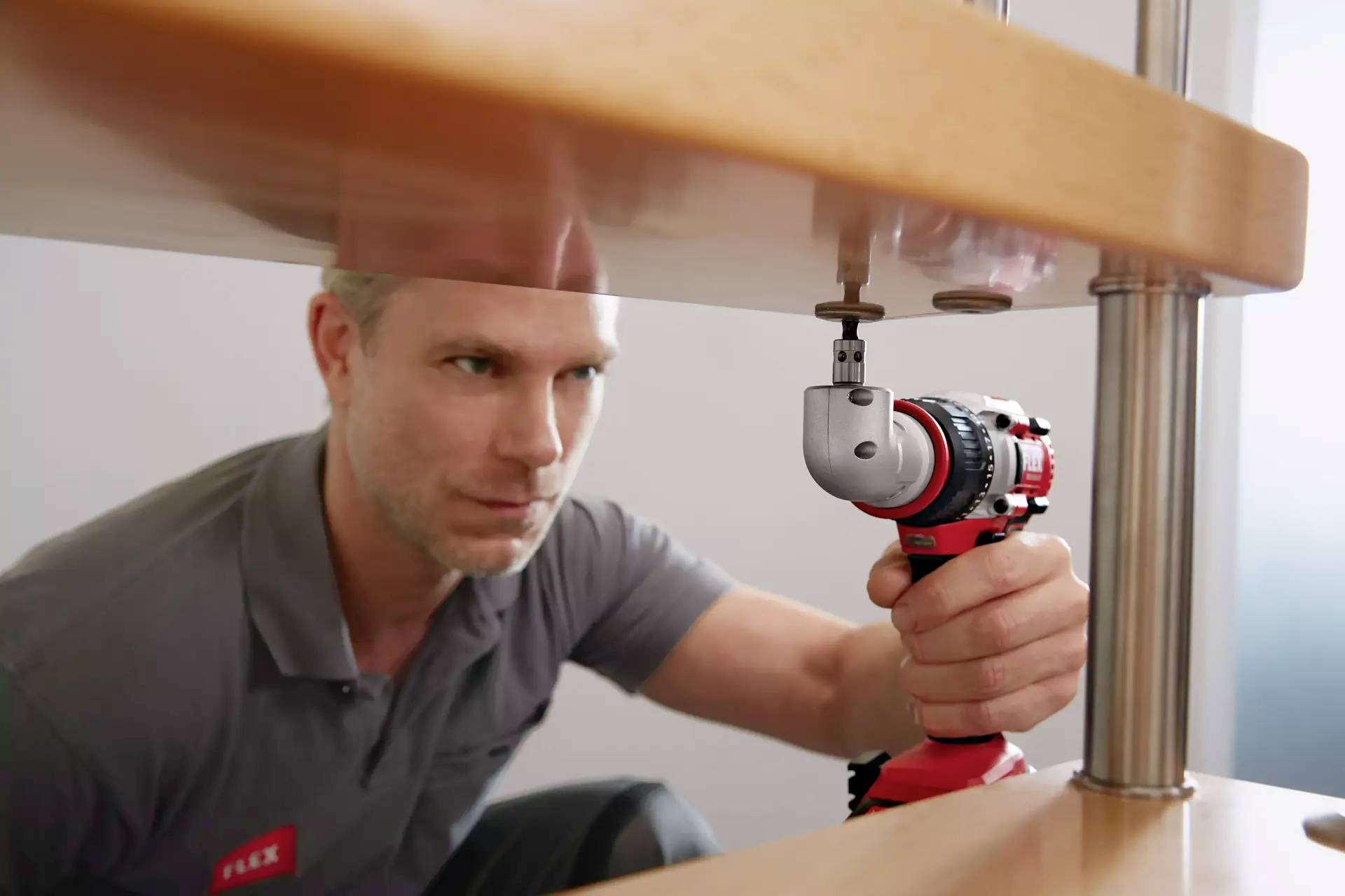 Mężczyzna w szarym polo używa akumulatorowego wkrętaka z elastycznym bitem, aby zamocować śrubę pod drewnianym stołem.
