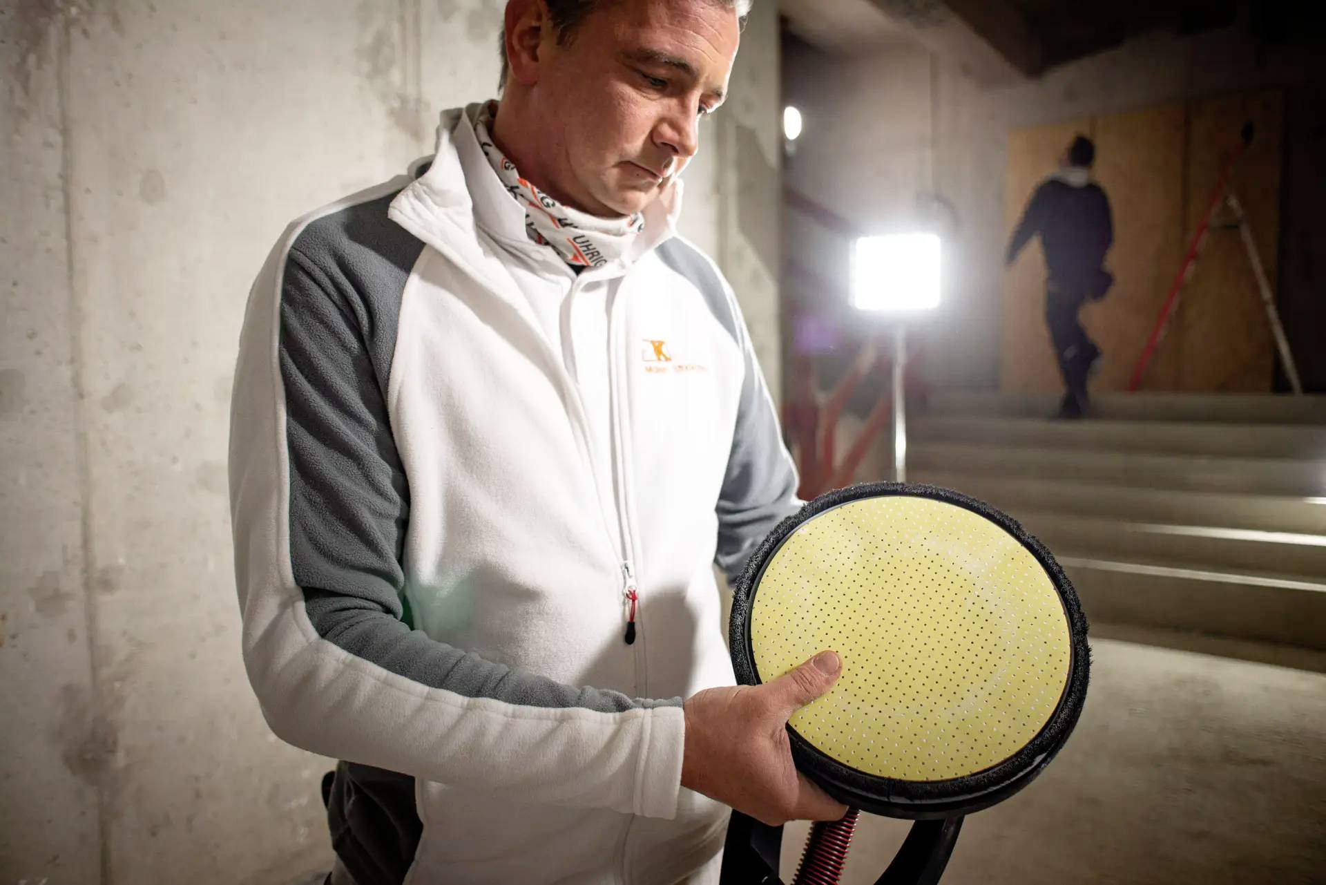 Mężczyzna w odzieży roboczej trzyma tarczę szlifierską w przemysłowym otoczeniu, podczas gdy w tle ktoś wspina się po schodach.