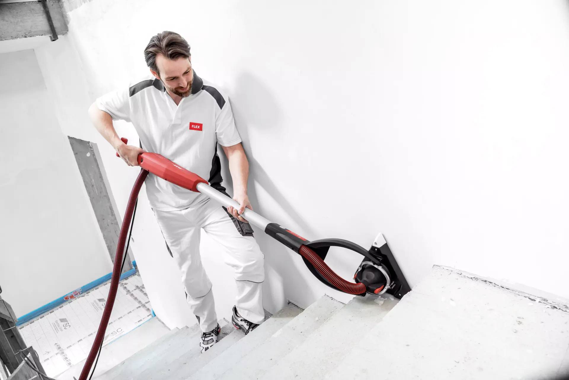 Robotnik odkurza schody przemysłowym odkurzaczem. Ubrany jest w strój roboczy i wydaje się znajdować w budynku będącym w trakcie budowy.