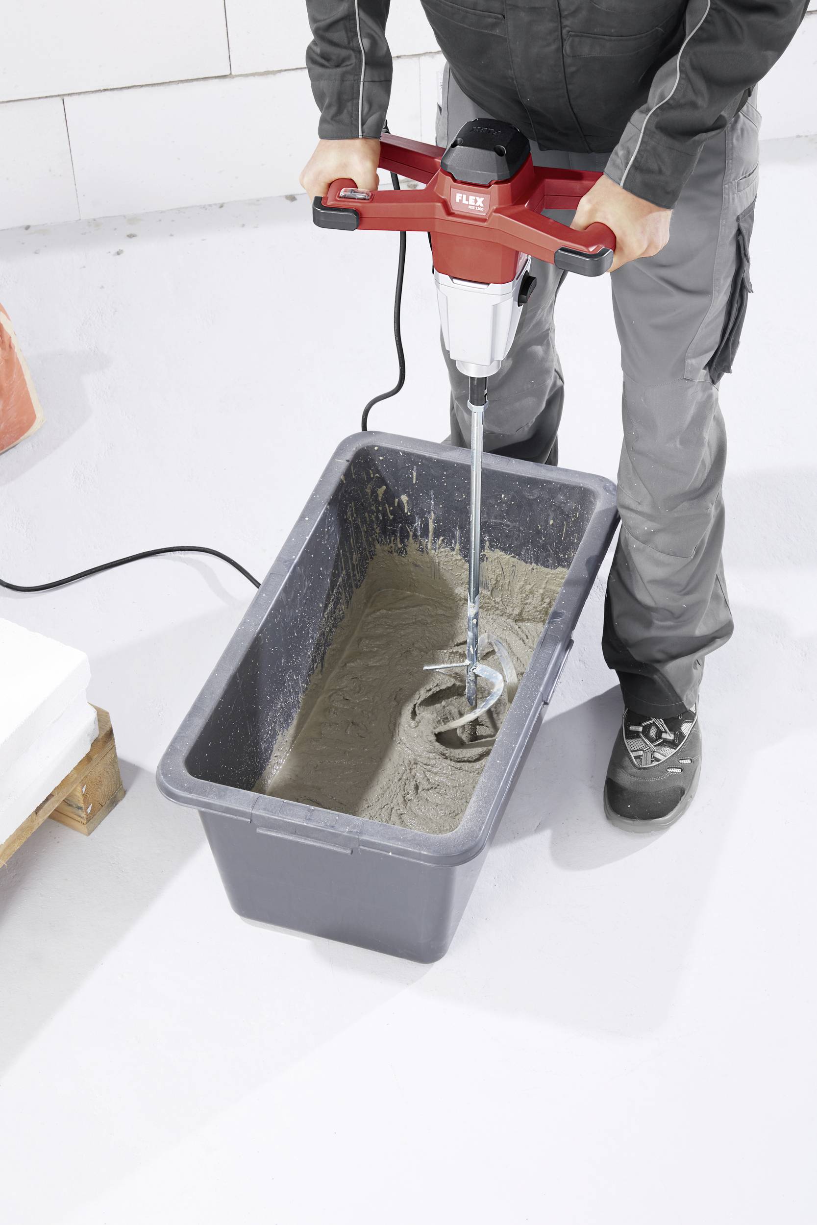 Osoba miesza cement lub beton elektrycznym mieszadłem w prostokątnym pojemniku.